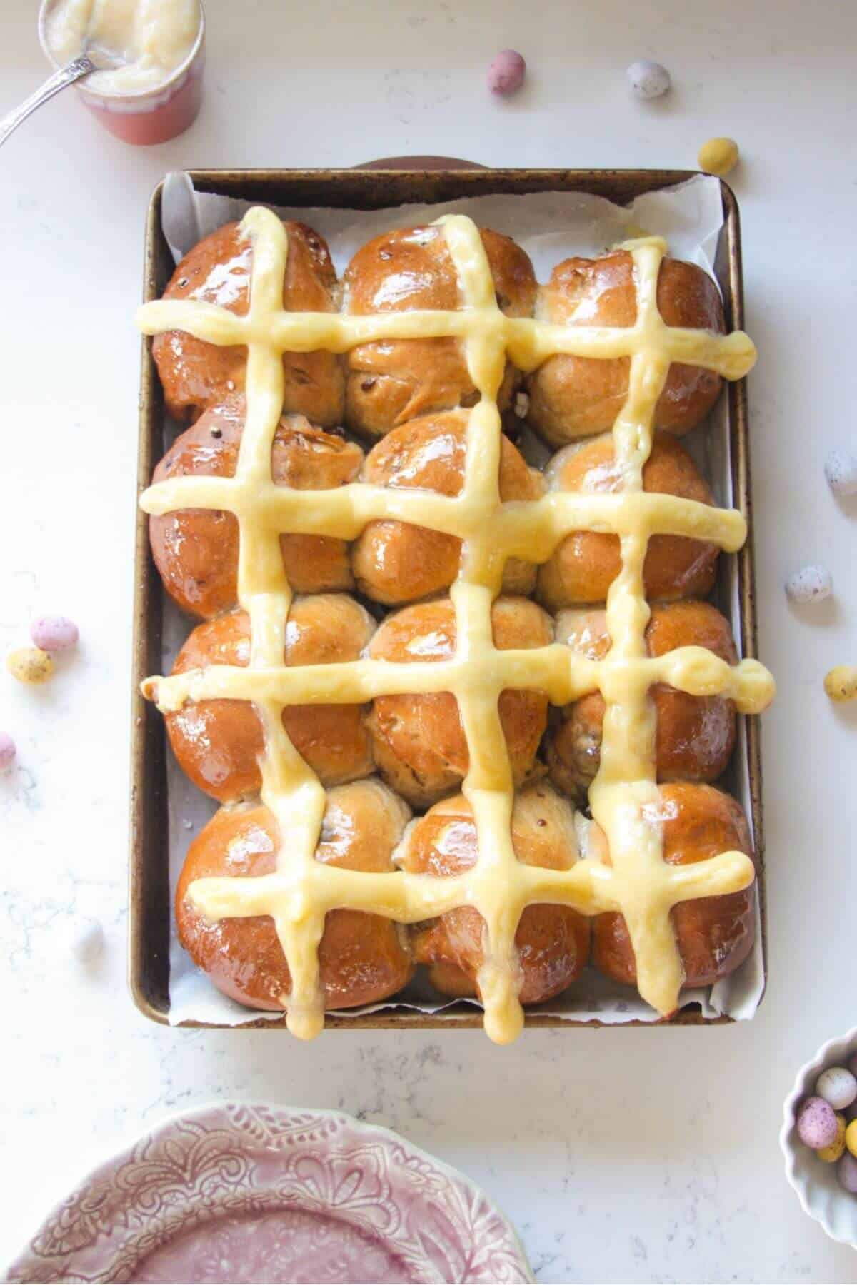 12 glazed mini egg chocolate hot cross buns in a lined oven tray with mini eggs on the bench next to them.