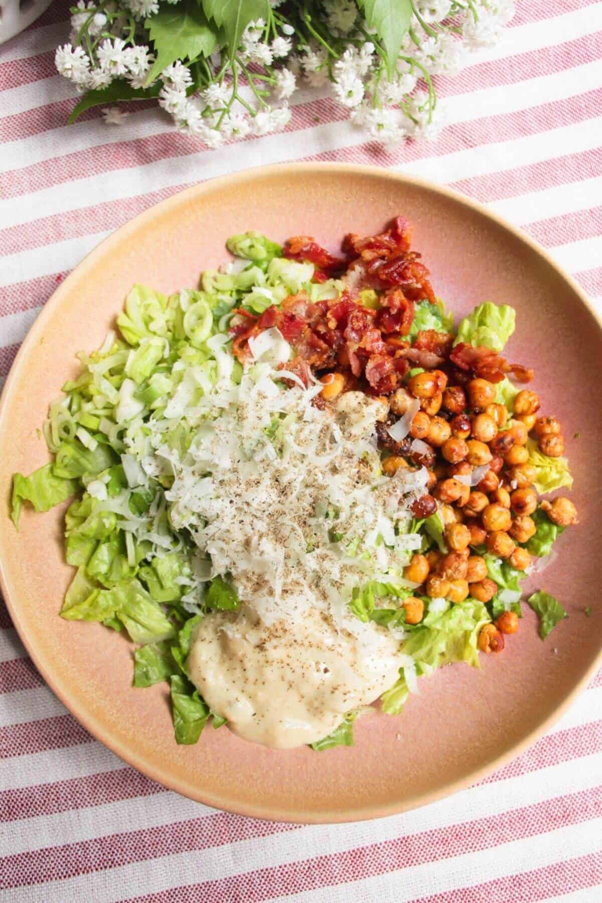 Chopped lettuce, scallions, bacon, parmesan, chickpeas and dressing in a pink bowl.