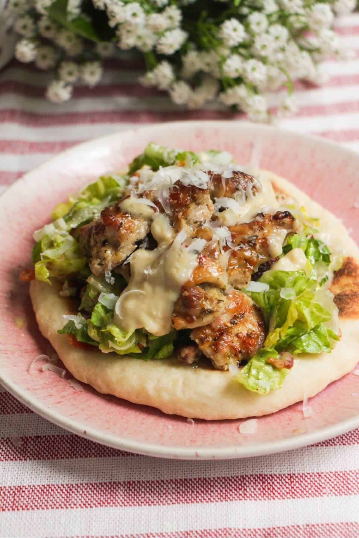 Flatbread on a plate topped with Caesar salad, charred chicken pieces and creamy Caesar dressing.