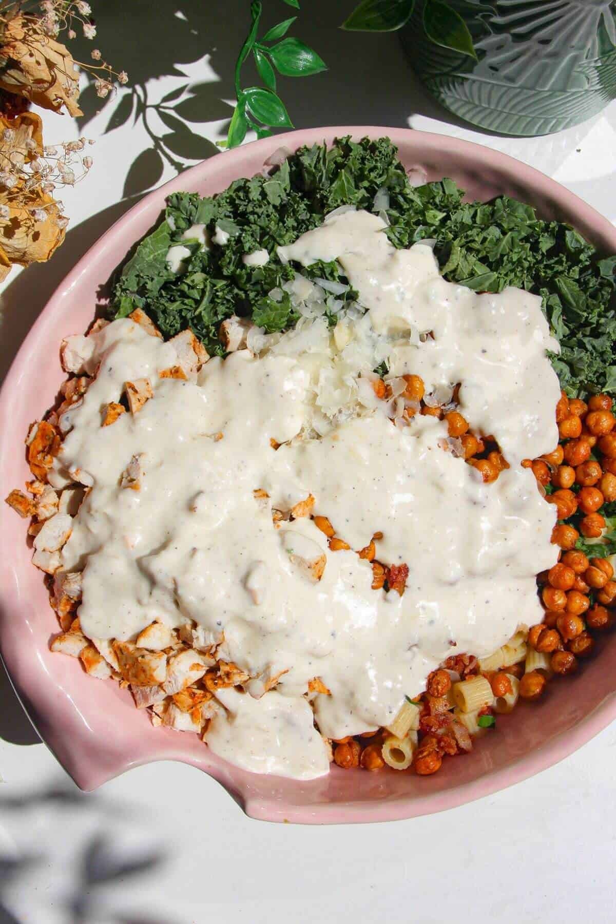 Caesar dressing poured over pasta salad in mixing bowl.