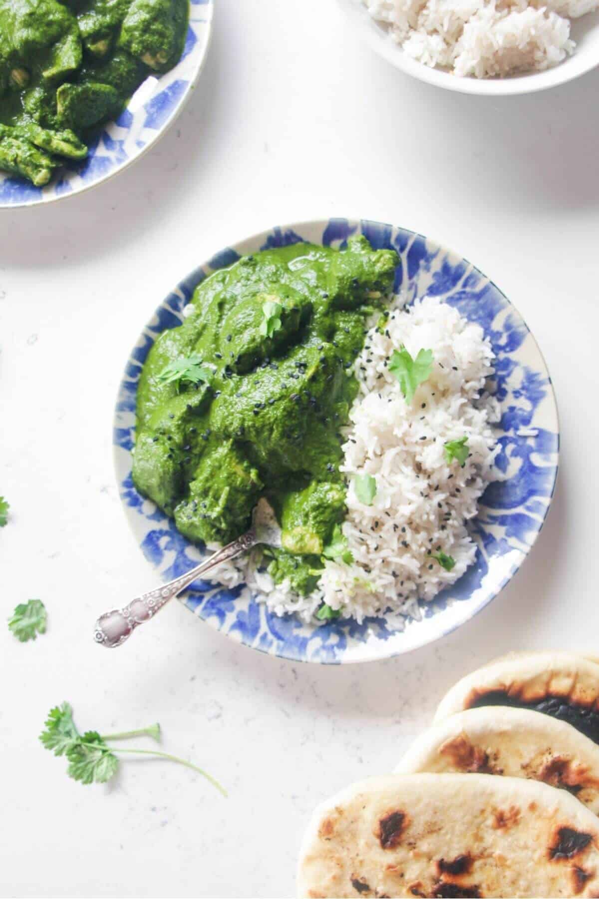 Chicken saag on a white and blue plate with rice, with more curry and naan on the side.