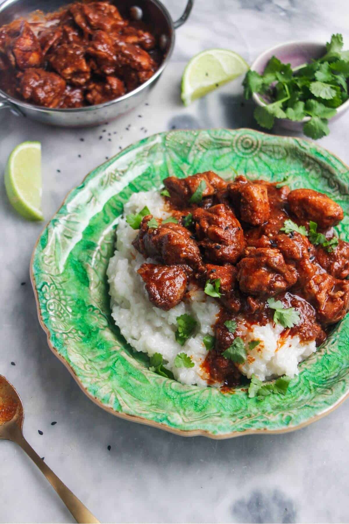 Close up of chicken rogan josh on rice on a small green plate with more curry in the background.