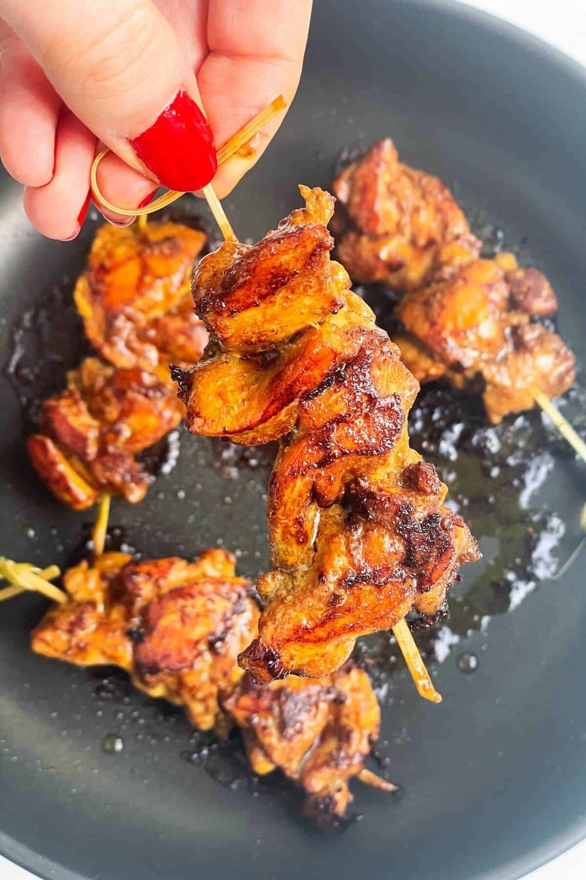 Hand holding a cooked chicken skewer with more chicken in the background.