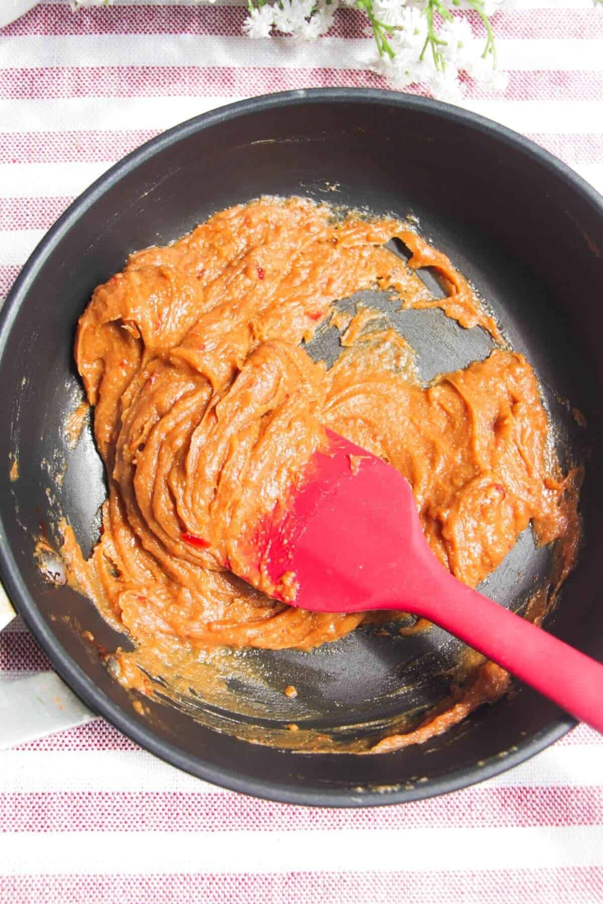 Red apatula stirring peanut sauce ingredients in a small pan.