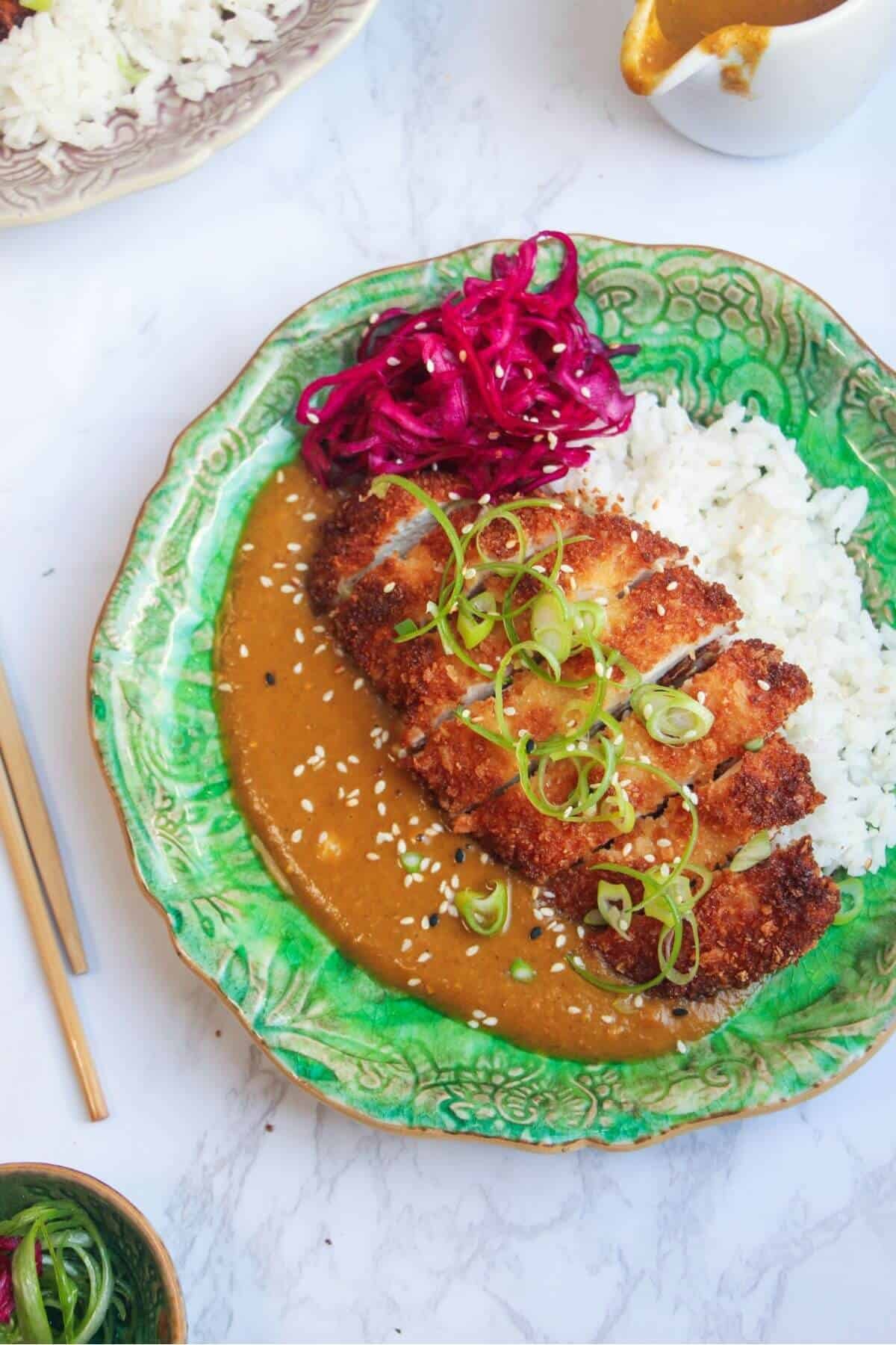 Sliced, breaded chicken breast on top of rice, curry sauce and pickled red cabbage on a green plate.