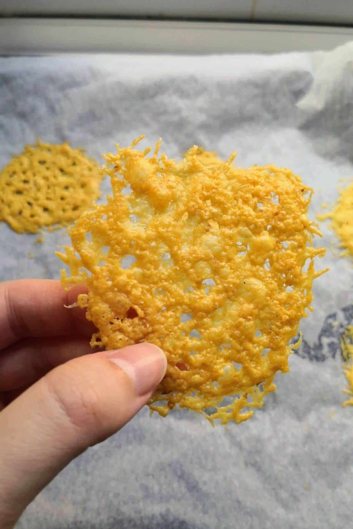 Hand holding up a parmesan crisp with more parmesan crisps on an oven tray in the background.
