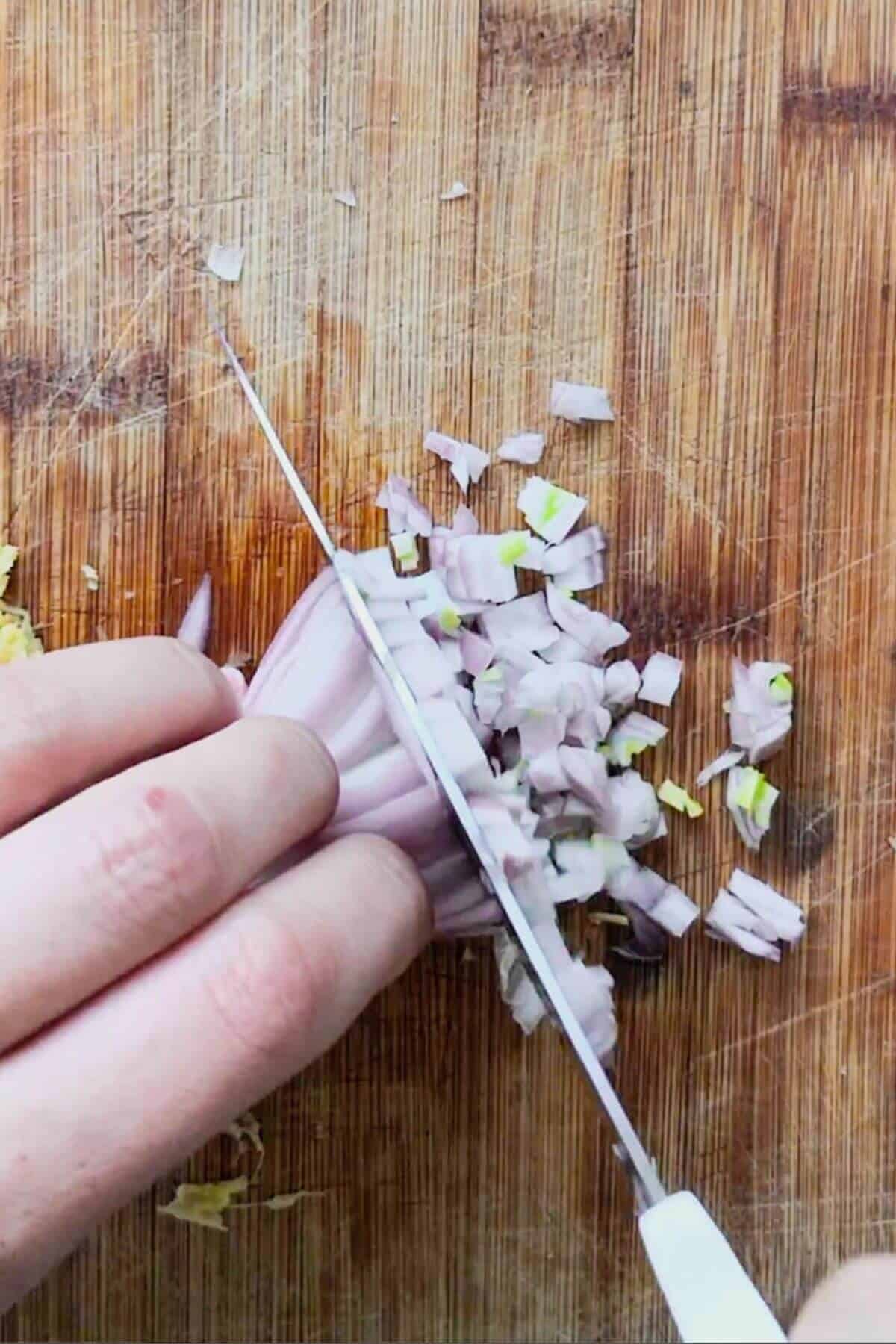 Hand with large knife chopping shallot on a wooden board.