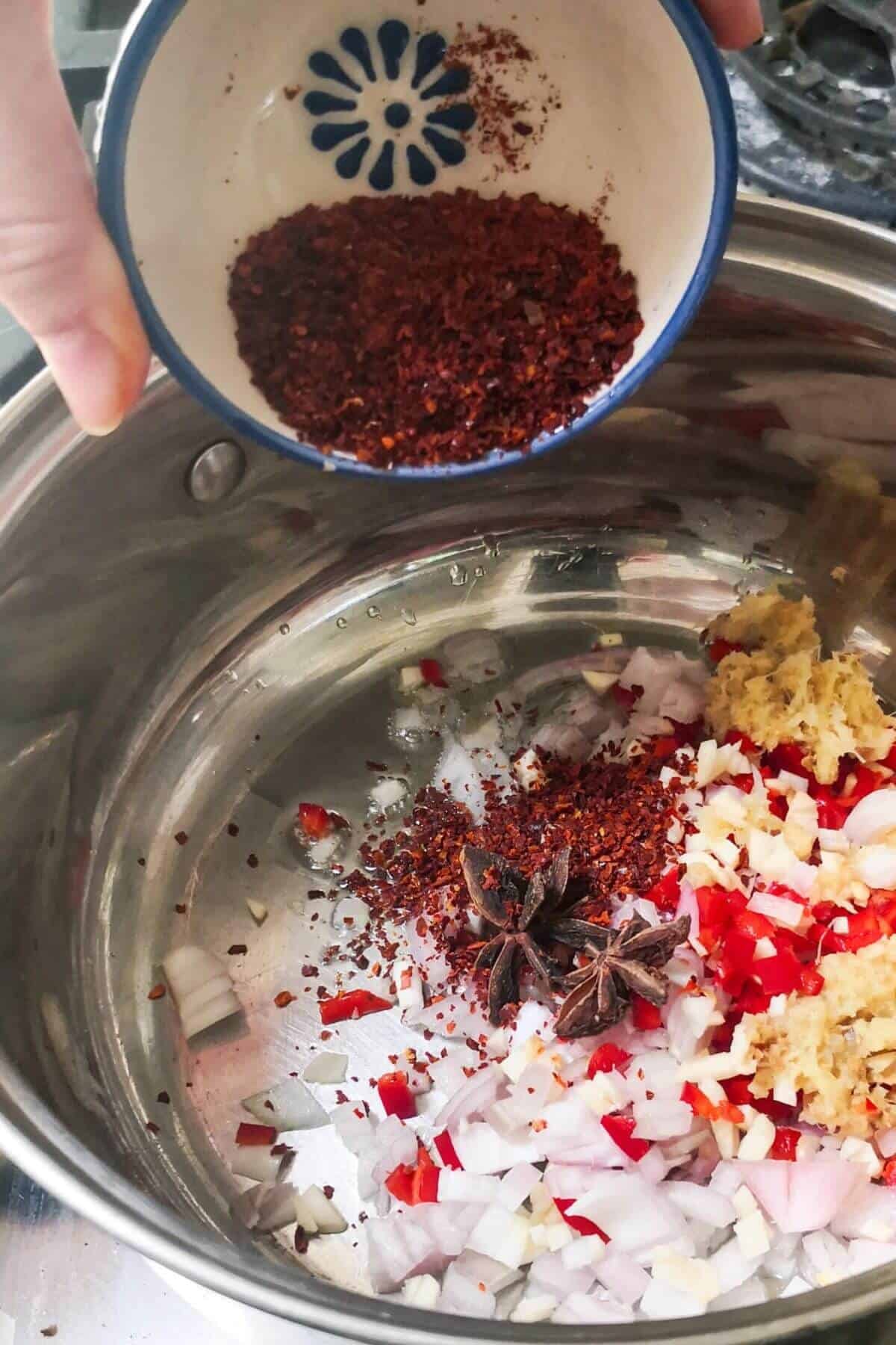 Spices being added to small silver pot with other chilli oil ingredients in a small silver pot.