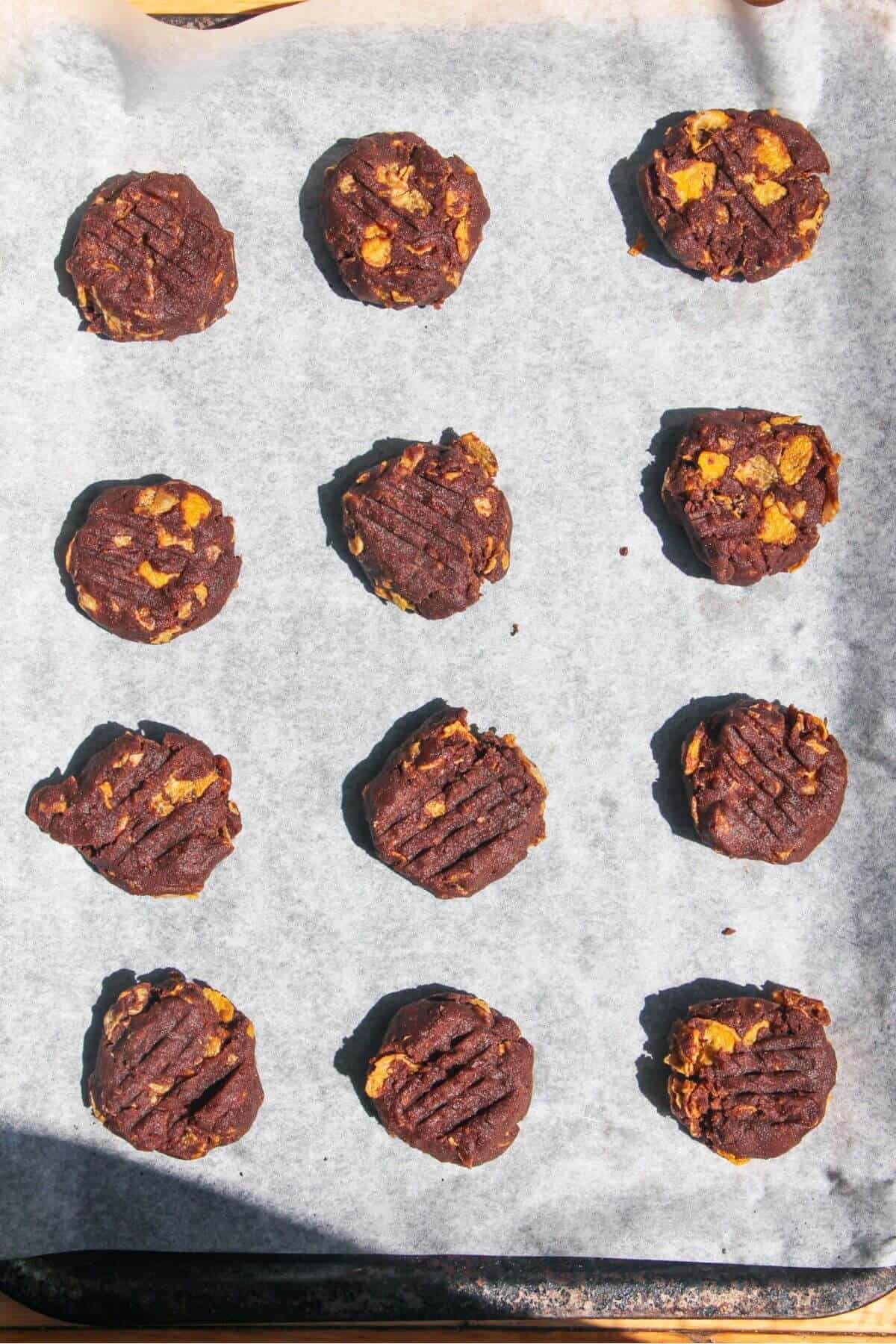 12 slightly flattened balls of chocolate Afghan biscuit dough on a lined baking tray.