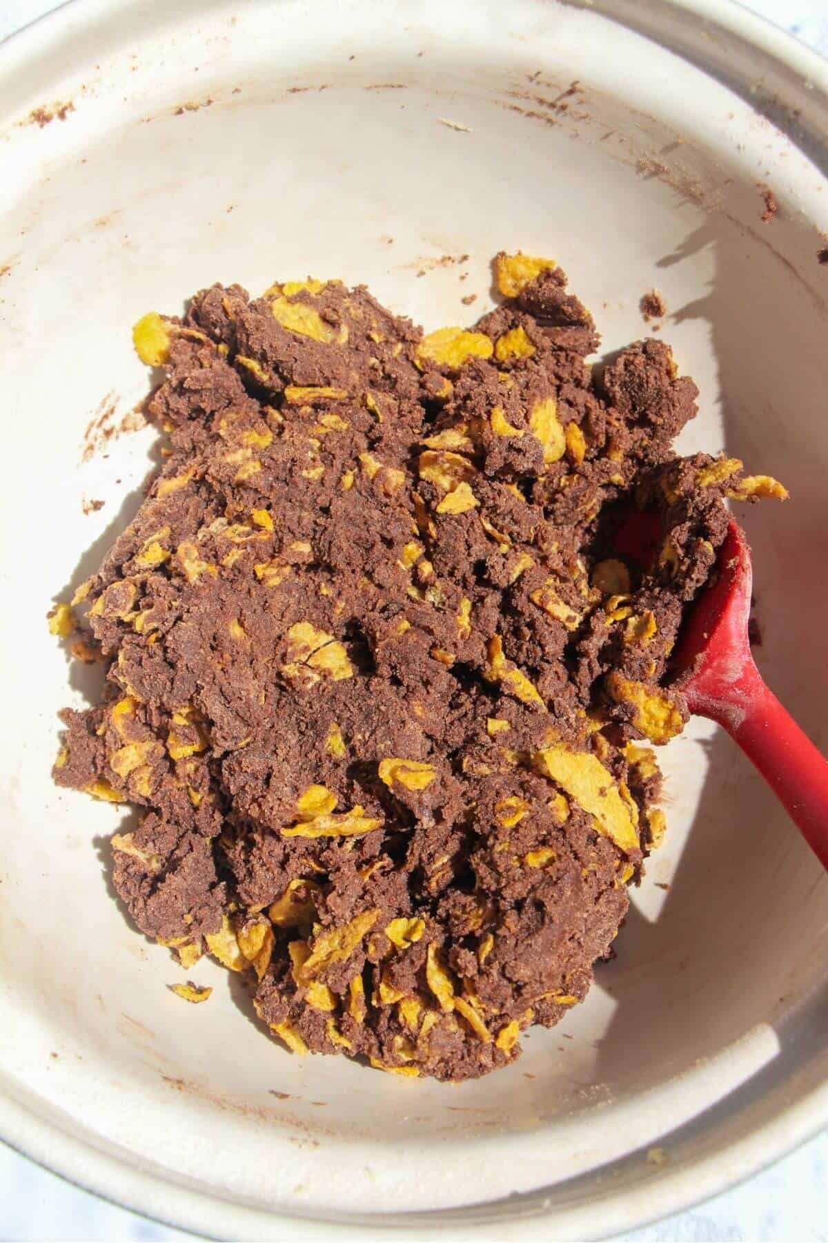 Cornflakes added and mixed into Afghan biscuit dough in a large white mixing bowl.