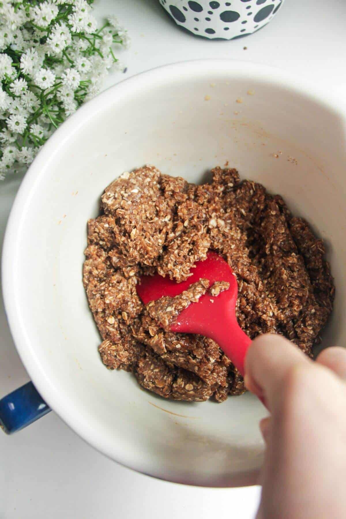 Red spatula stirring Anzac biscuit dough in a mixing bowl.