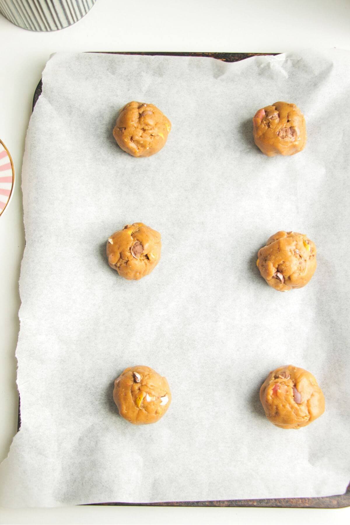 Six cookie dough balls on a baking paper lined oven tray.