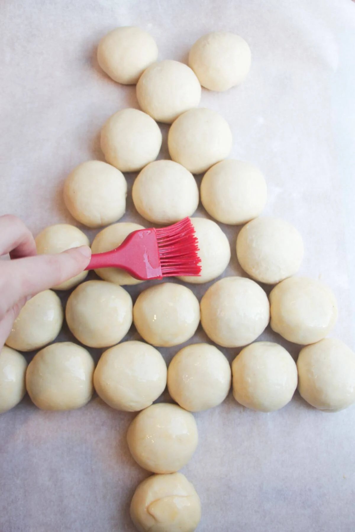 Hand holding a red brush, egg-washing the Christmas tree dough balls.