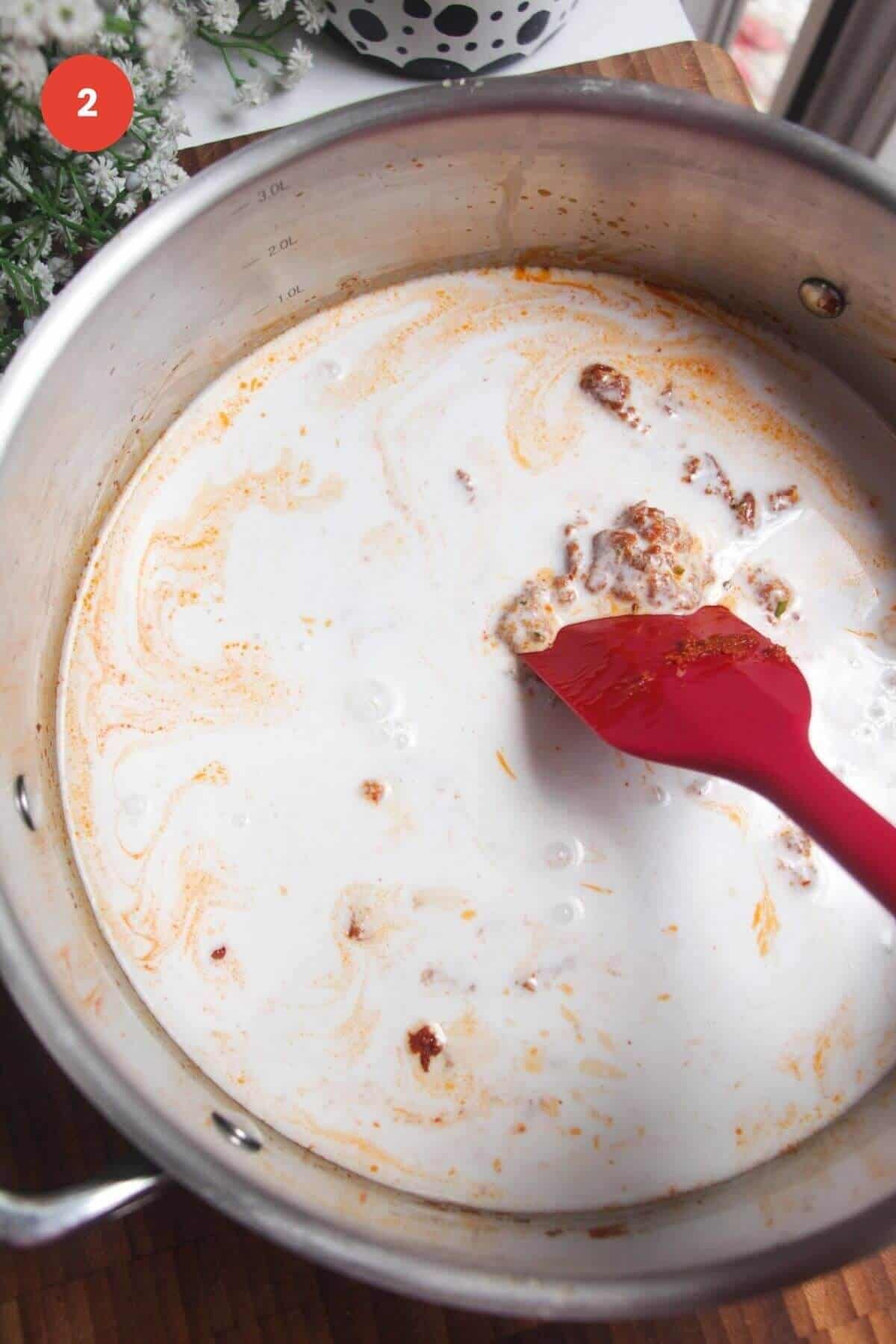 Spatula stirring coconut milk through red curry paste in a large pan.