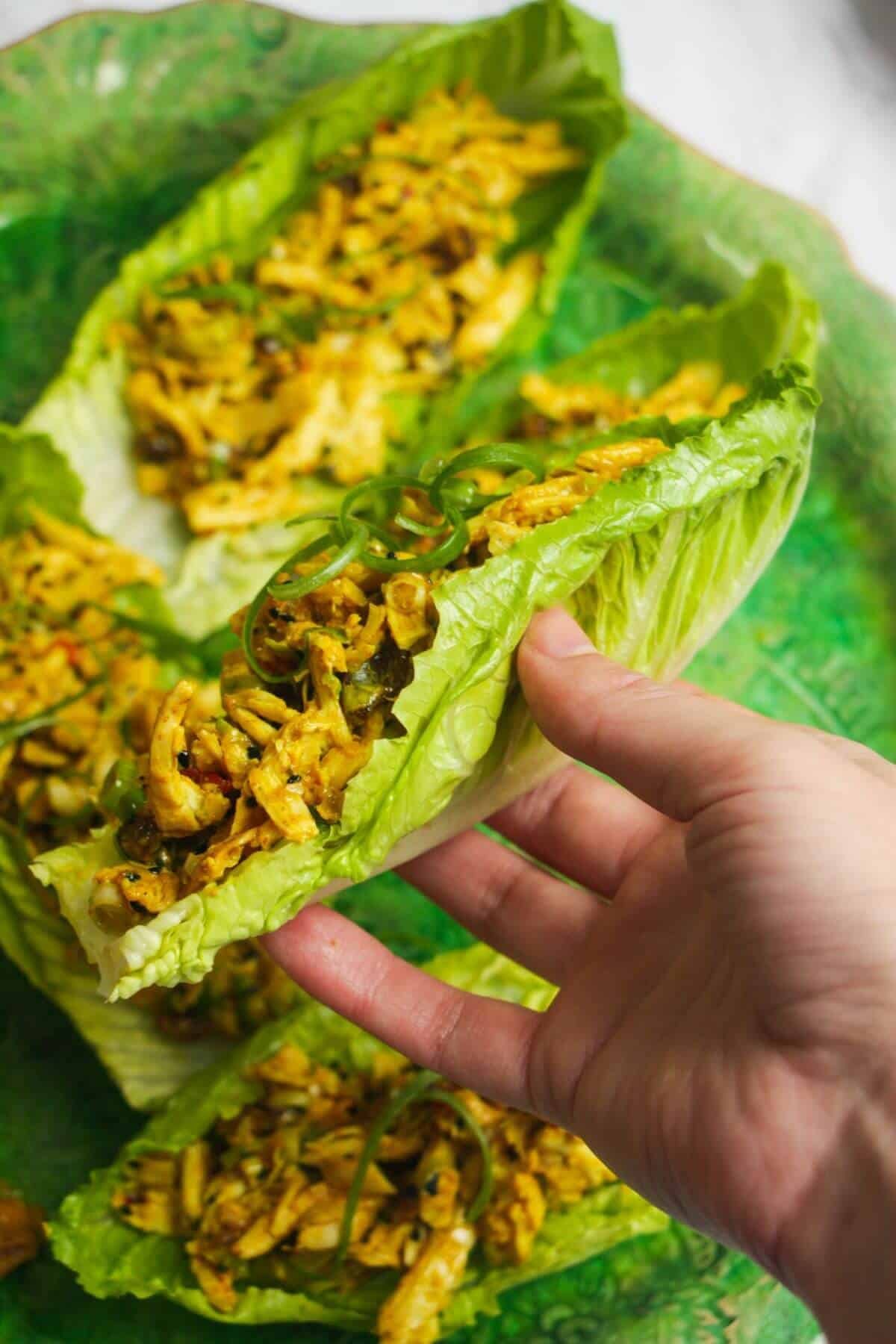 Hand holding a lettuce leaf with coronation chicken inside with more coronation chicken in the background.