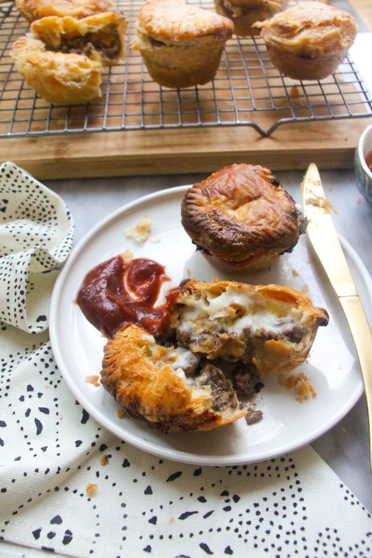 One cut open mince and cheese pie on a plate with beef and cheese filling oozing out, with tomato sauce on the side and another small mince and cheese pie on the plate behind it.