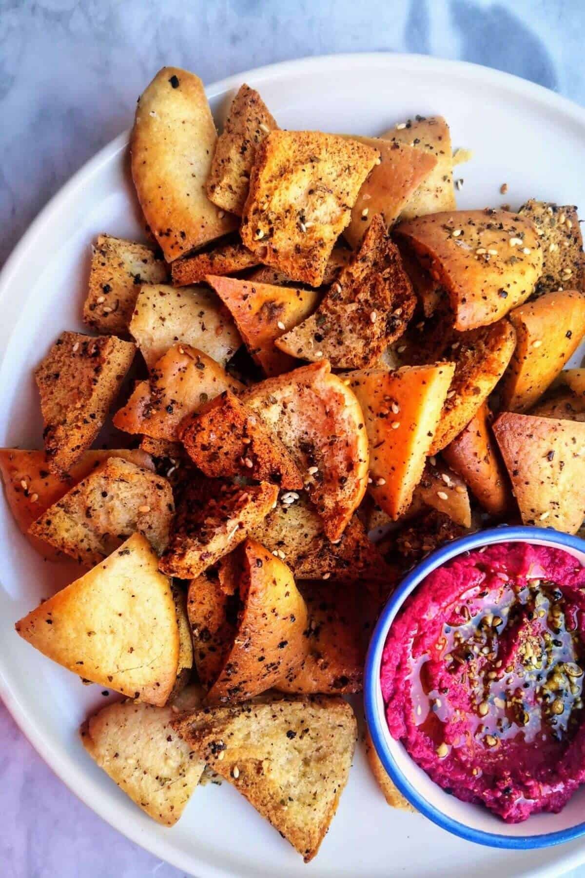 Pile of homemade pita chips on a white plate with a small bowl of beetroot hummus on the side.