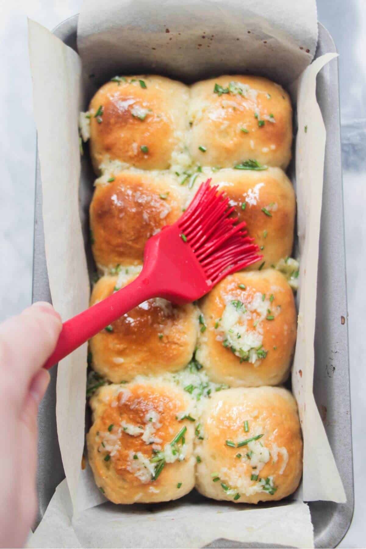 Garlic butter being brushed on 8 golden dinner rolls in a baking paper lined tin with a red brush.