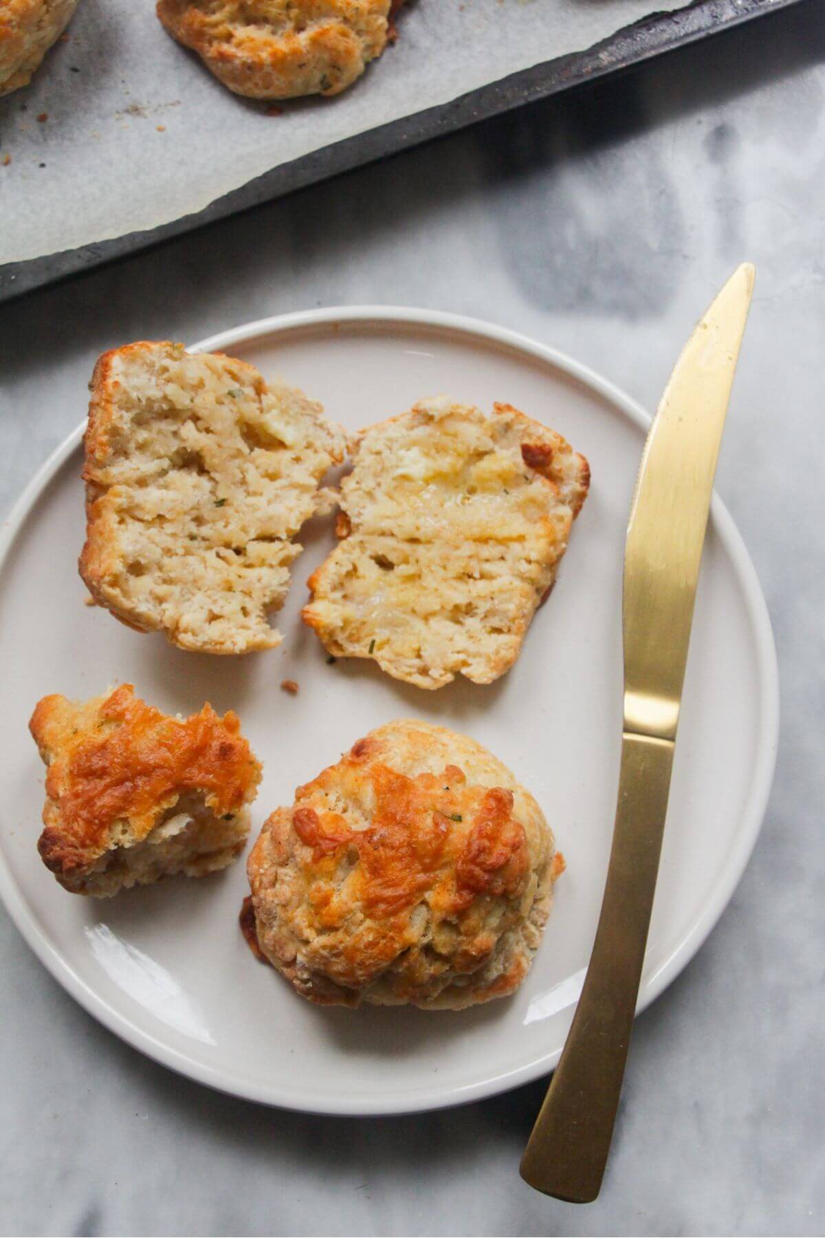 Small white plate with a whole and a sliced open cheese scone, with a gold knife on the side and tray of scones in the background.