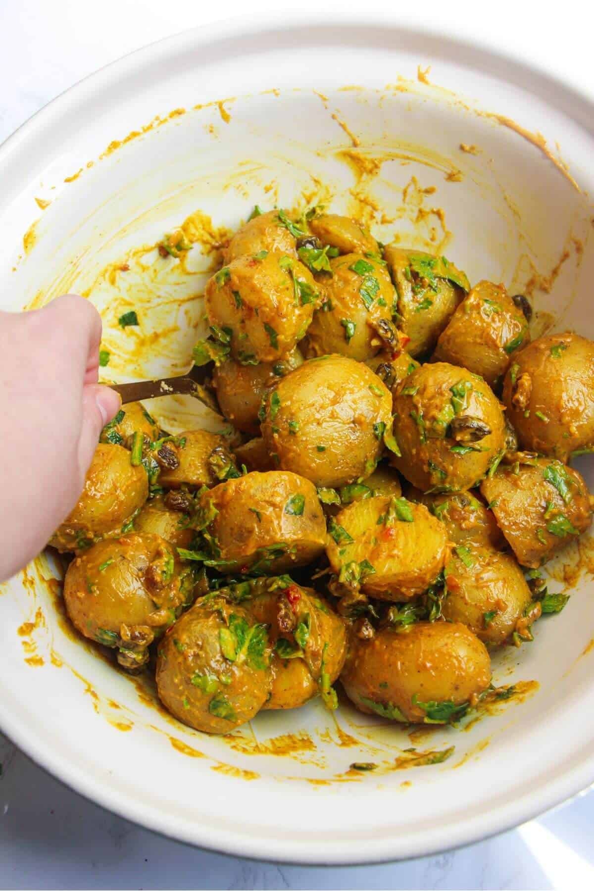 Hand holding a spoon mixing coronation potato salad in a large ceramic bowl.
