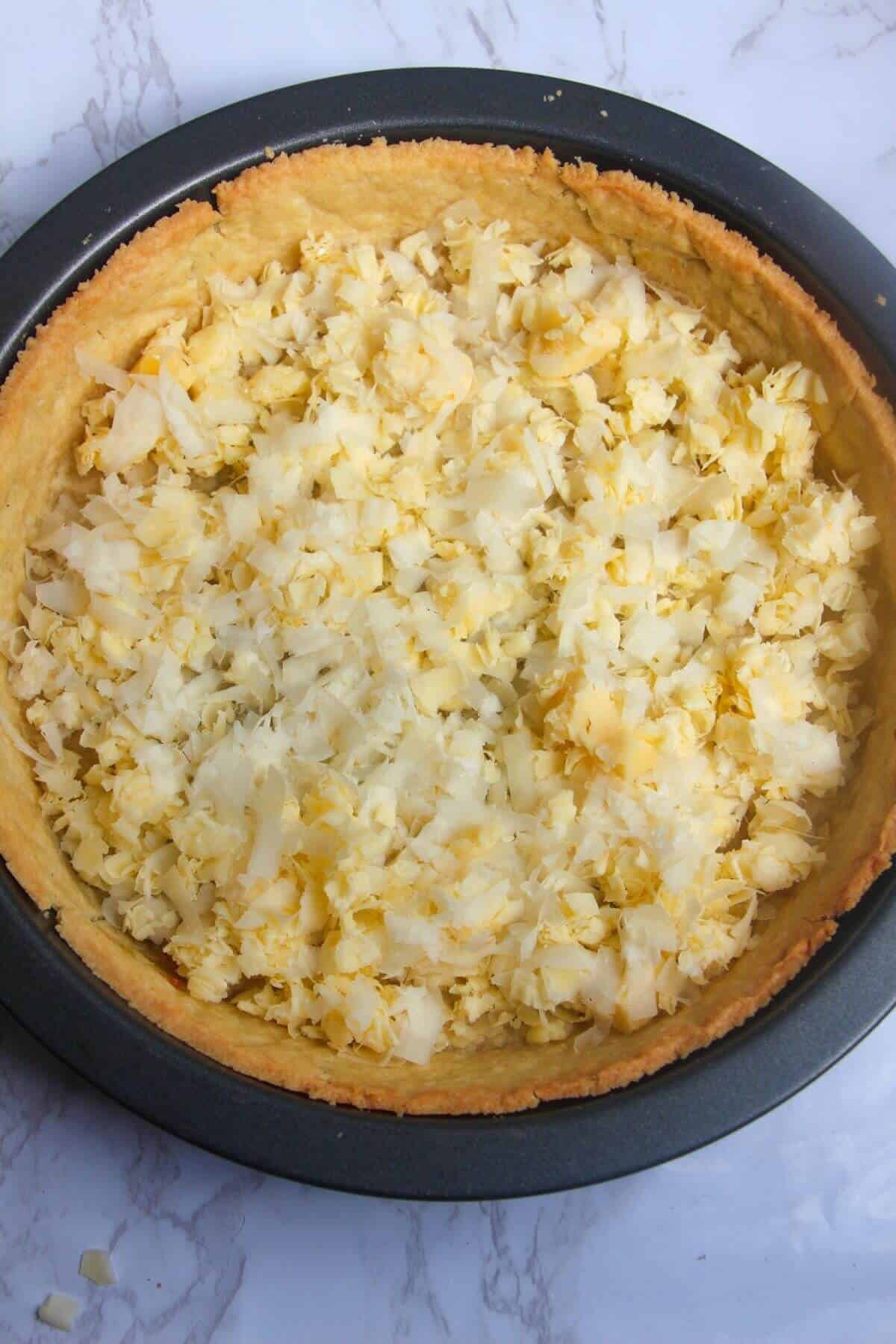Grated cheese scattered on the base of blind-baked pastry case in a tin.