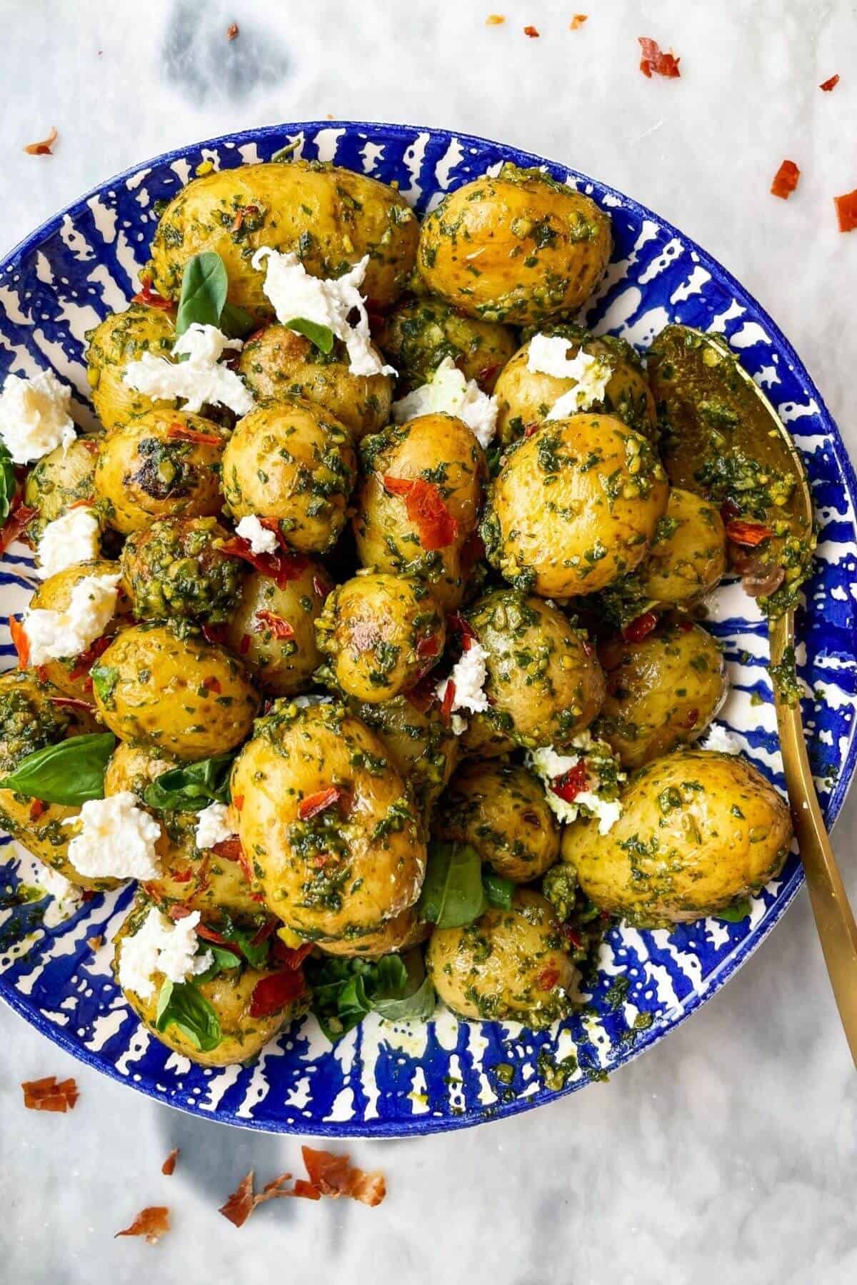 Pesto potatoes with crispy prosciutto and burrata in a blue and white bowl with a gold spoon on a grey marble background.