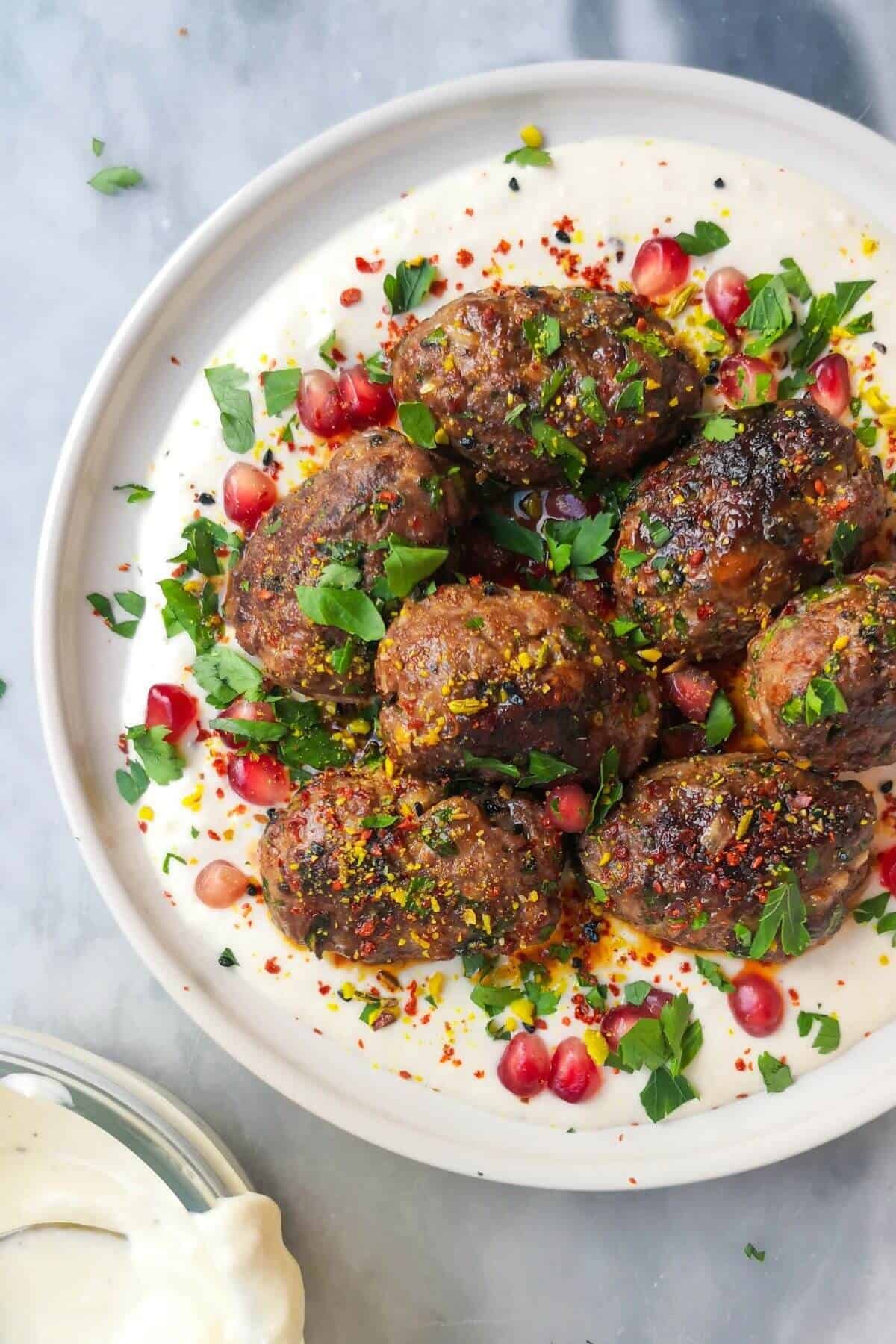 Lamb kofta with parsley, pomegranate seeds and dukkah on a white plate with yogurt tahini sauce, with a bowl of yogurt tahini sauce on the side.