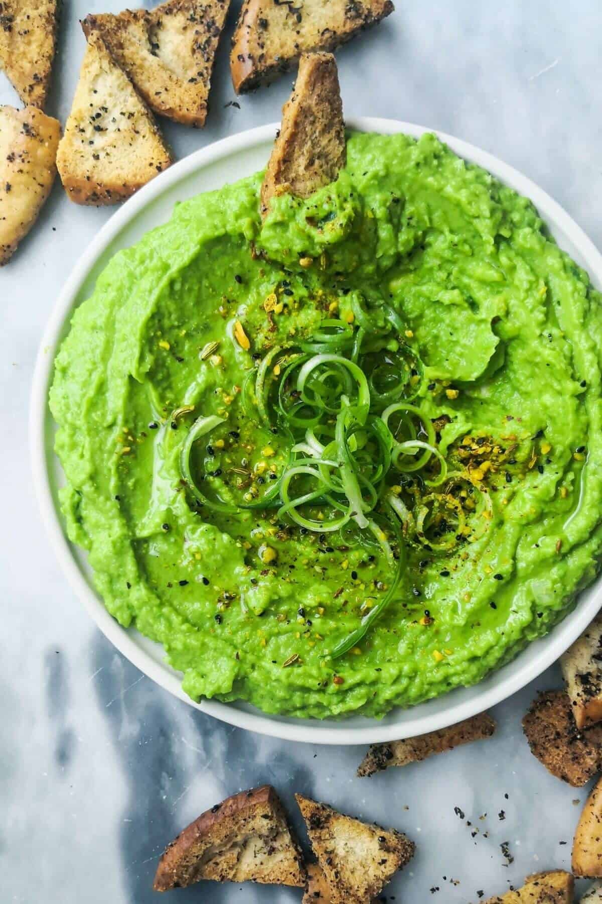 Pea hummus with avocado in a small white plate, pita chips on the side on a grey marble background