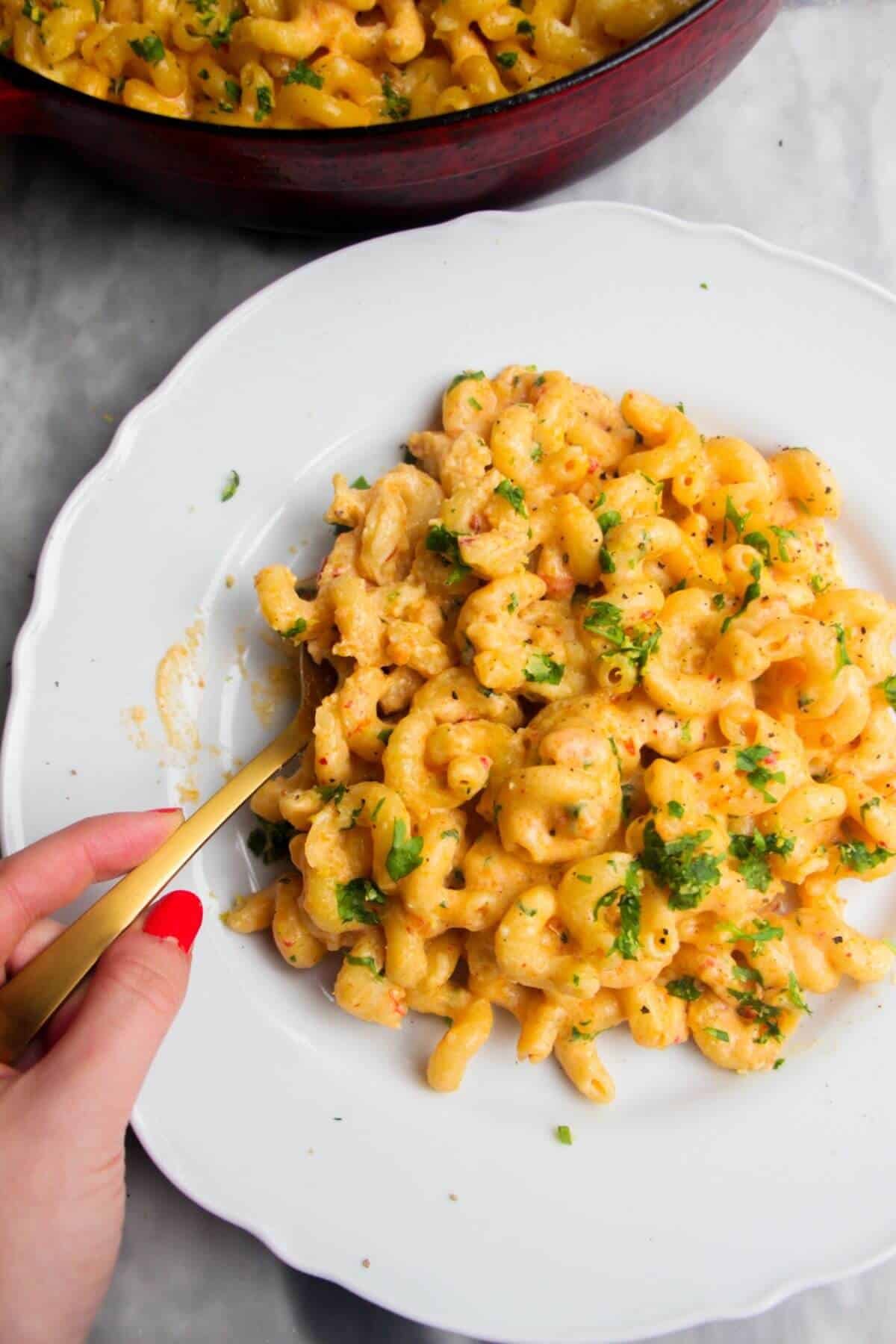 Spicy mac and cheese on a white plate with a hand holding a gold spoon, with more mac and cheese in the background.