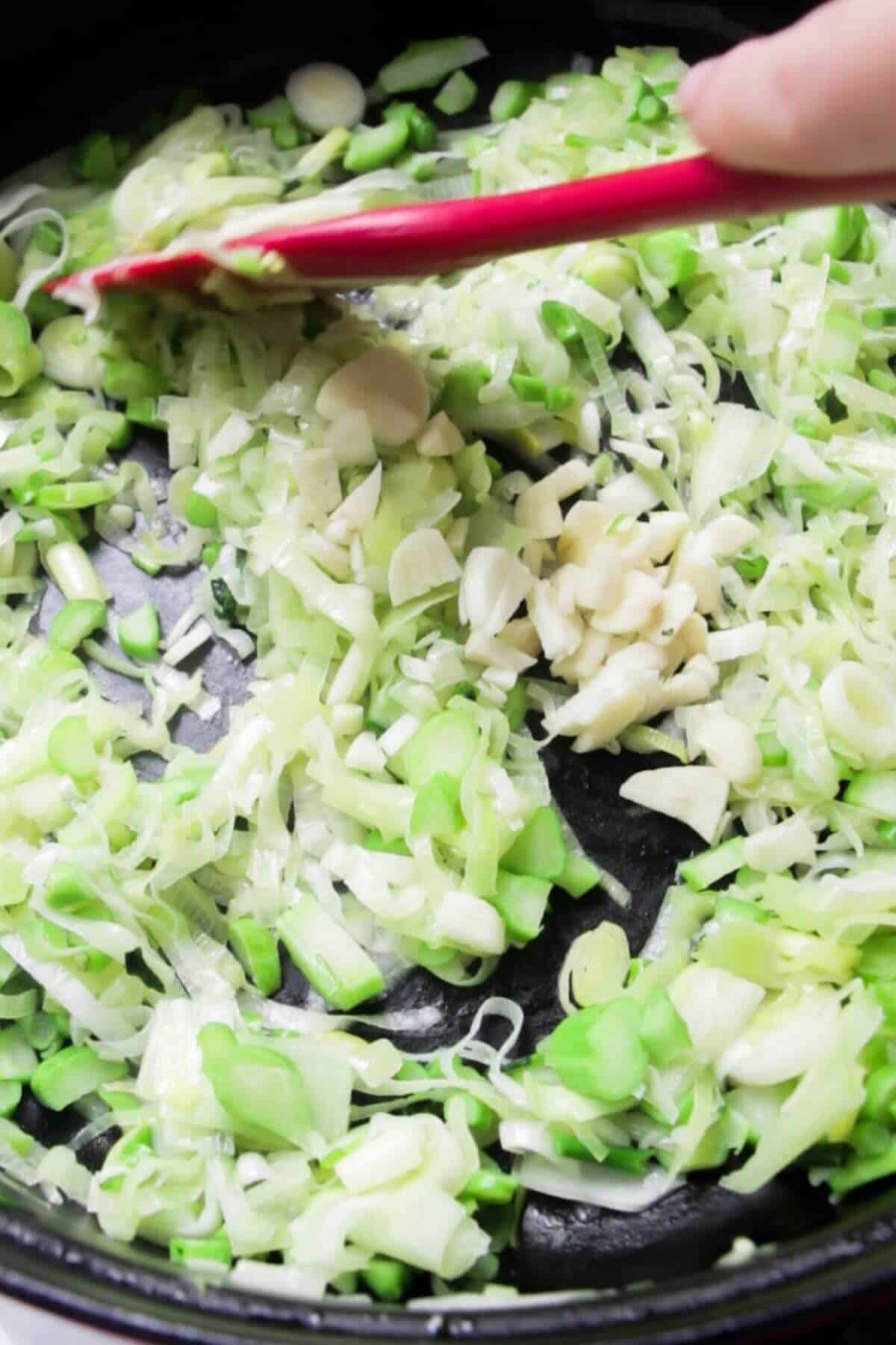 Red spatula stirring garlic, leeks and broccoli stems.