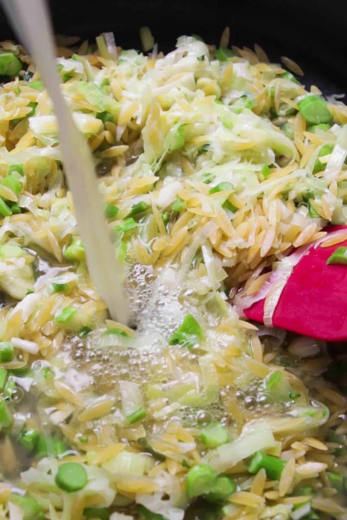 Stock being poured into a pan with orzo, leeks, garlic and broccolini stems.