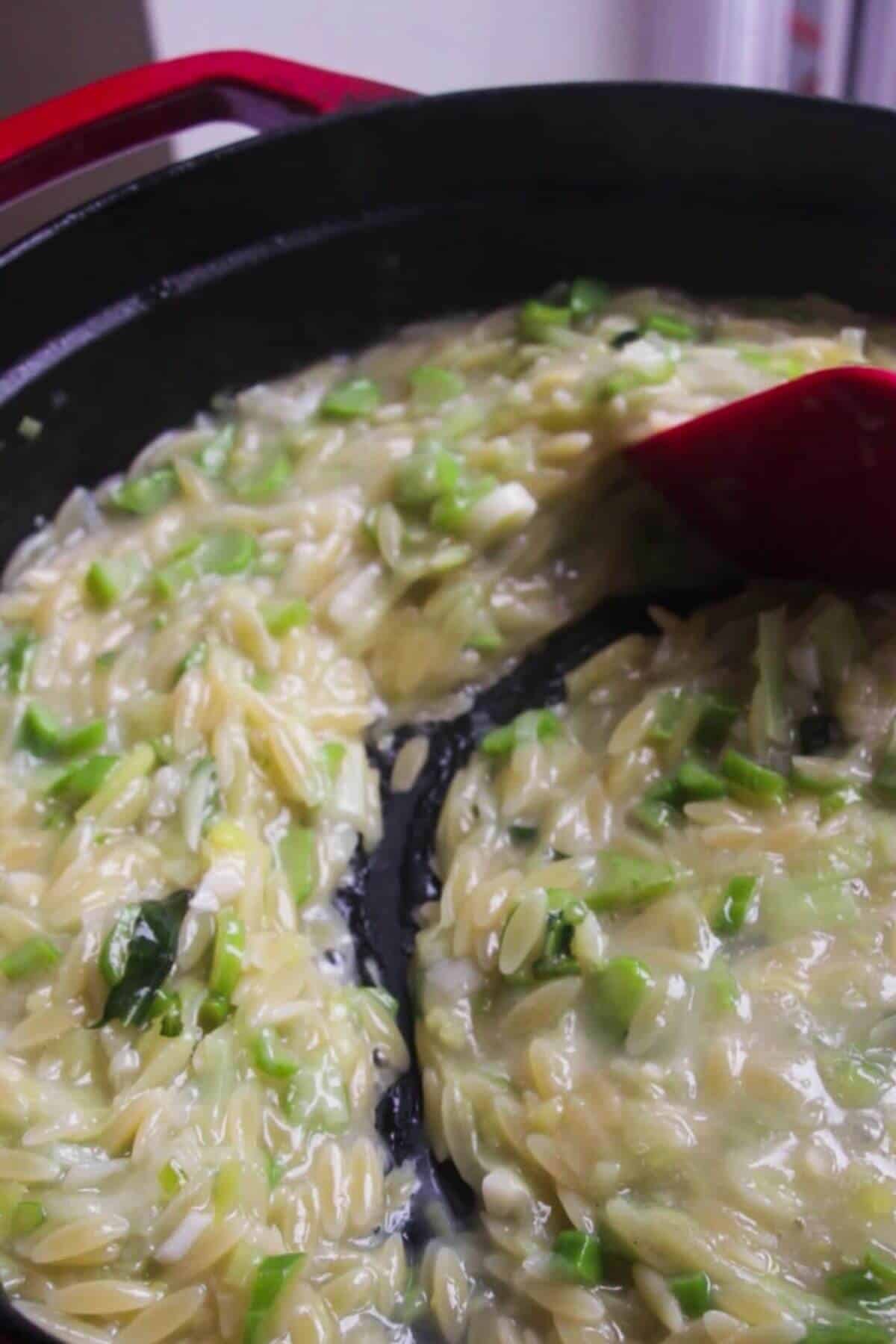 Red spatula stirring stock through creamy orzo in black and red pan.