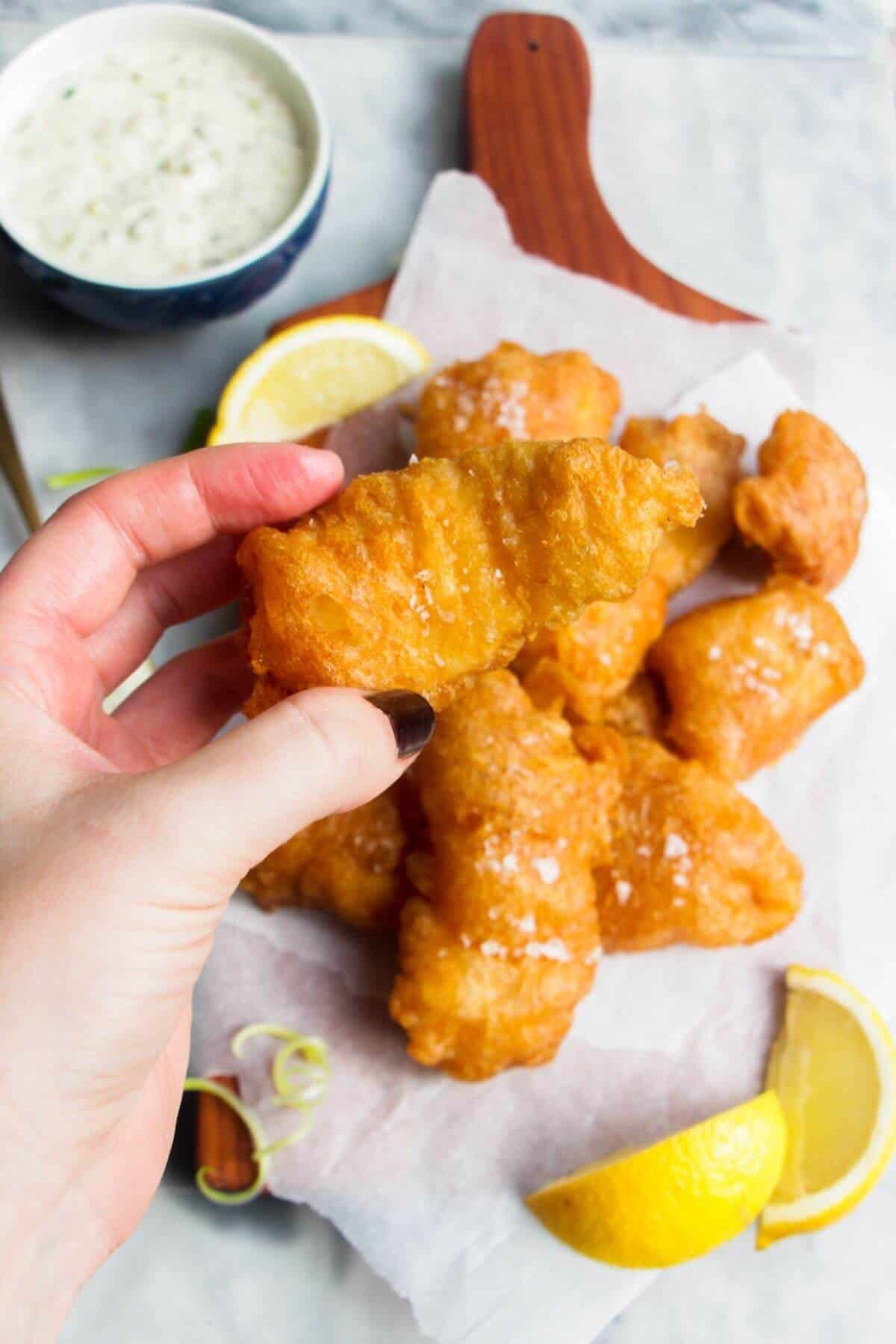 Hand holding a piece of golden beer battered fish with more fish in the background.