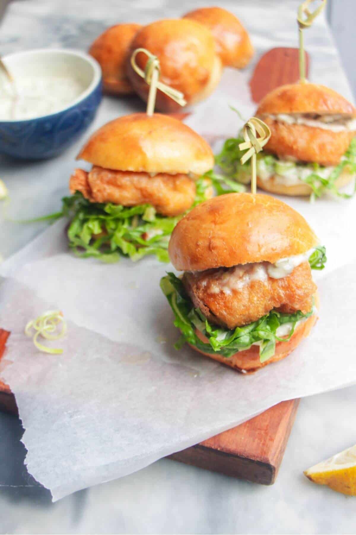 Three beer battered fish sliders on a baking paper lined wooden board with tartare sauce on the side.