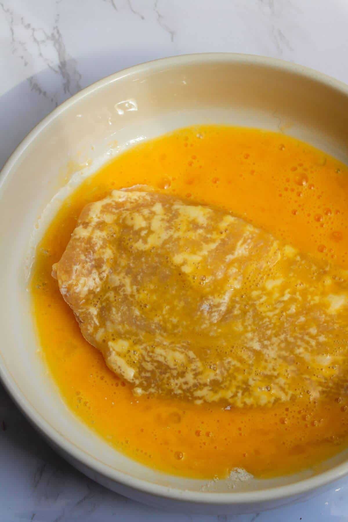 Large chicken fillet covered in whisked egg in a small white bowl.