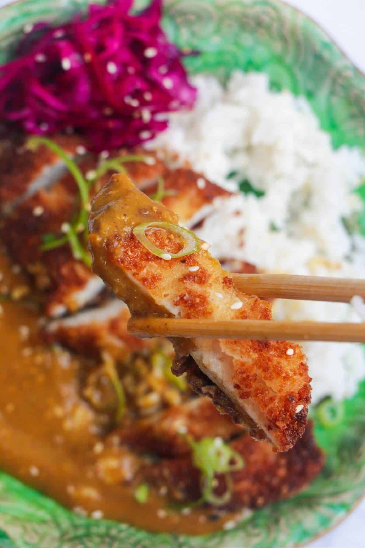 Chopsticks holding up a piece of breaded chicken with chicken katsu curry on a green plate in the background.