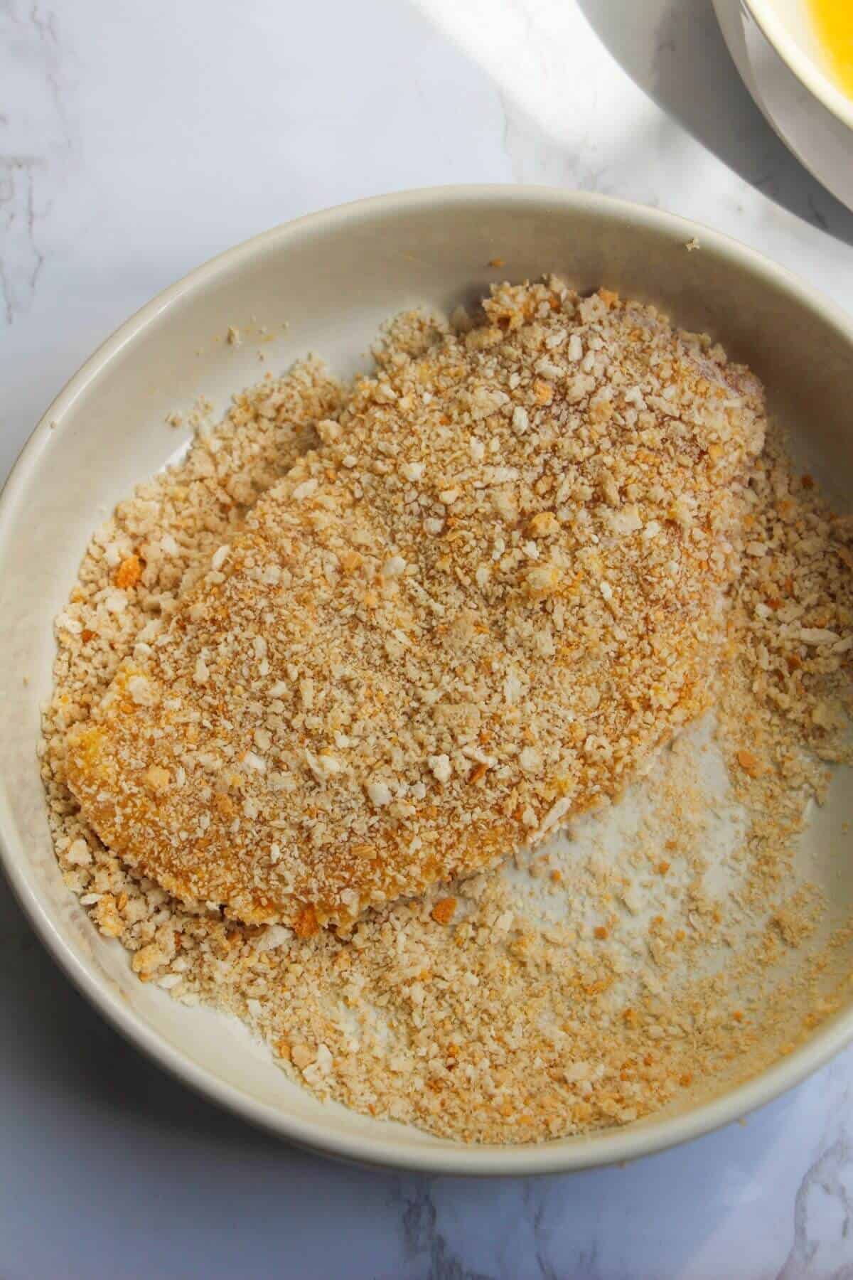 Large chicken breast fillet covered in breadcrumbs in a small white bowl.