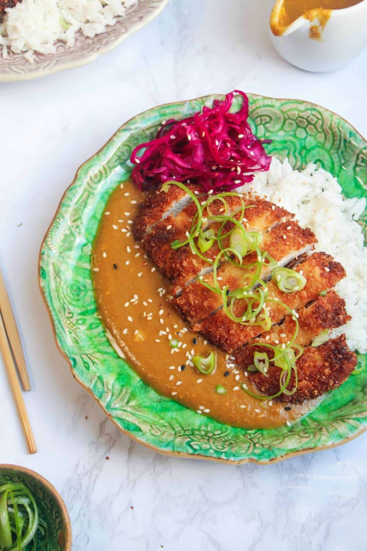 Sliced, breaded chicken breast on top of rice, curry sauce and pickled red cabbage on a green plate.