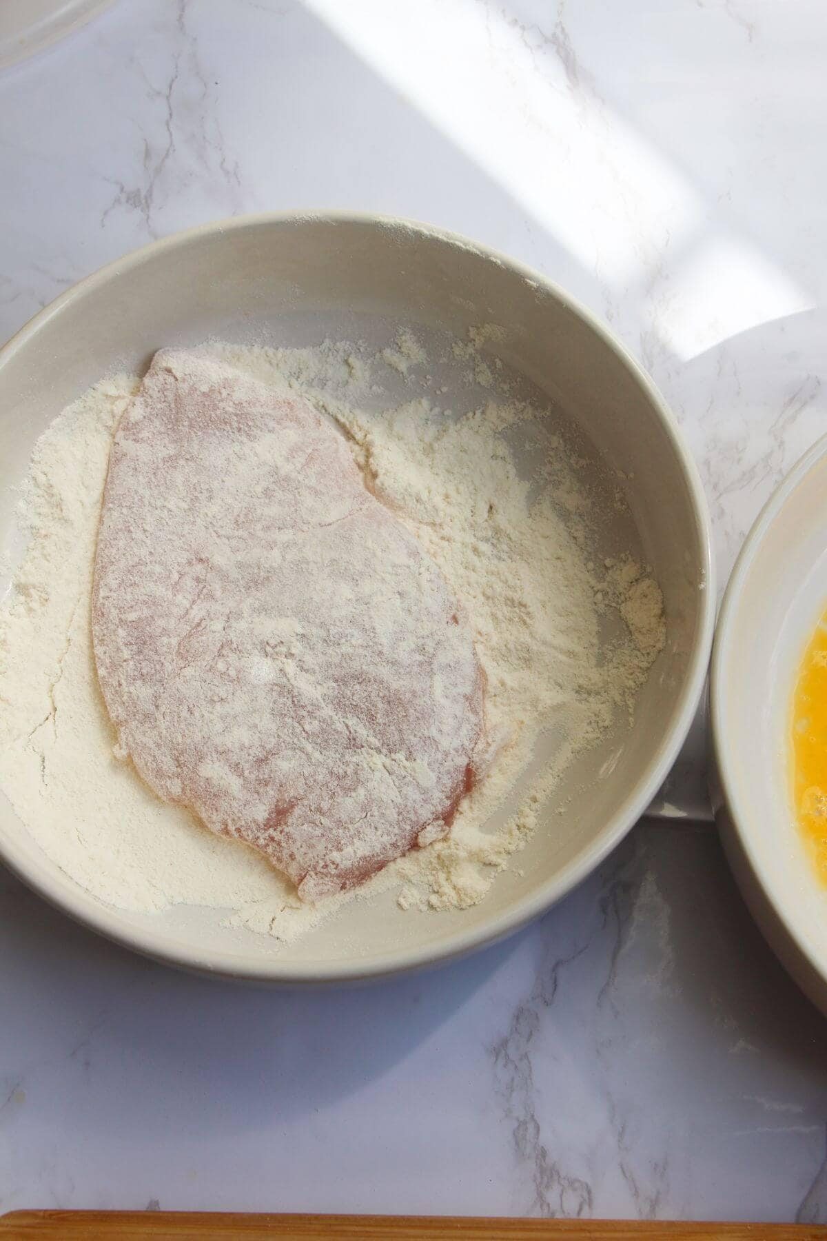 Large chicken fillet covered in flour in a small white bowl.