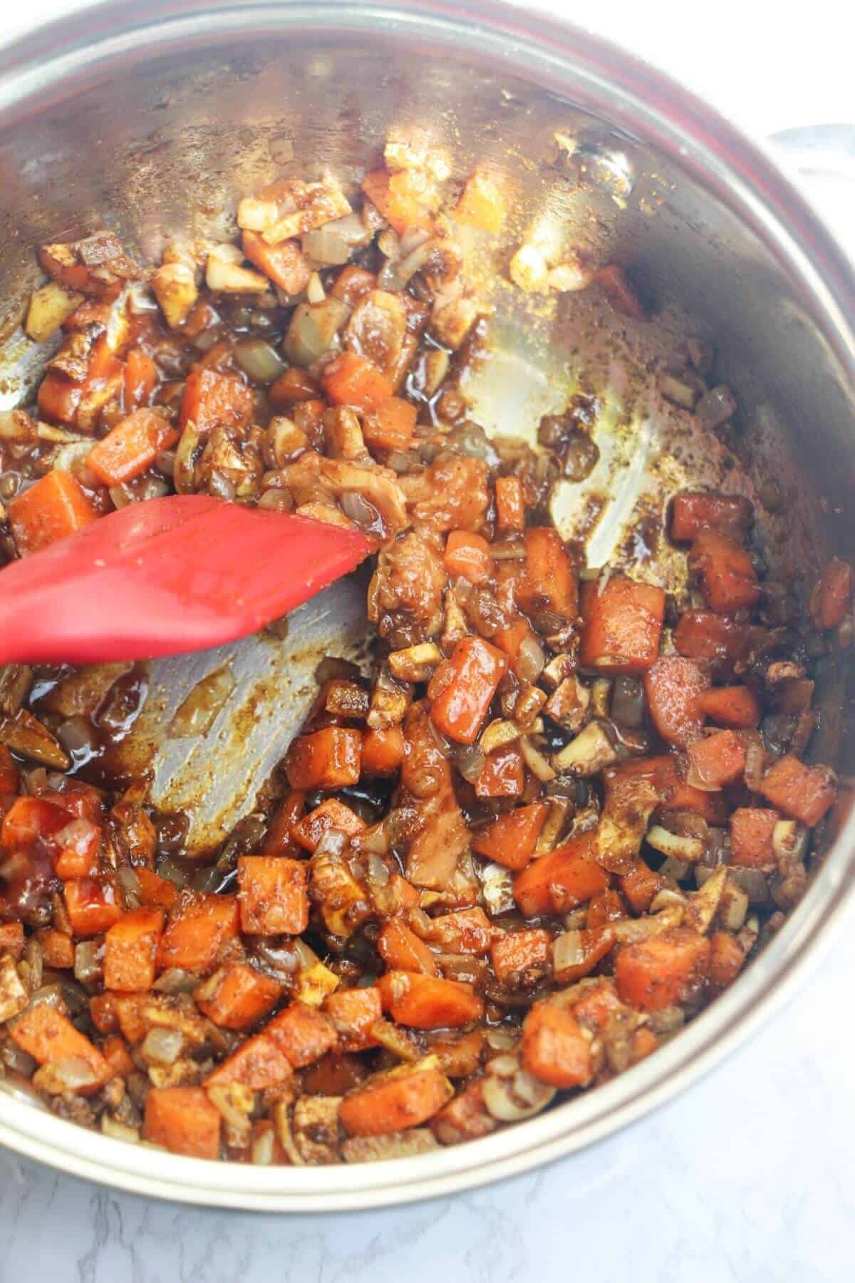 Red spatula stirring diced onion, carrots, ginger, garlic and spices in a small silver pot.