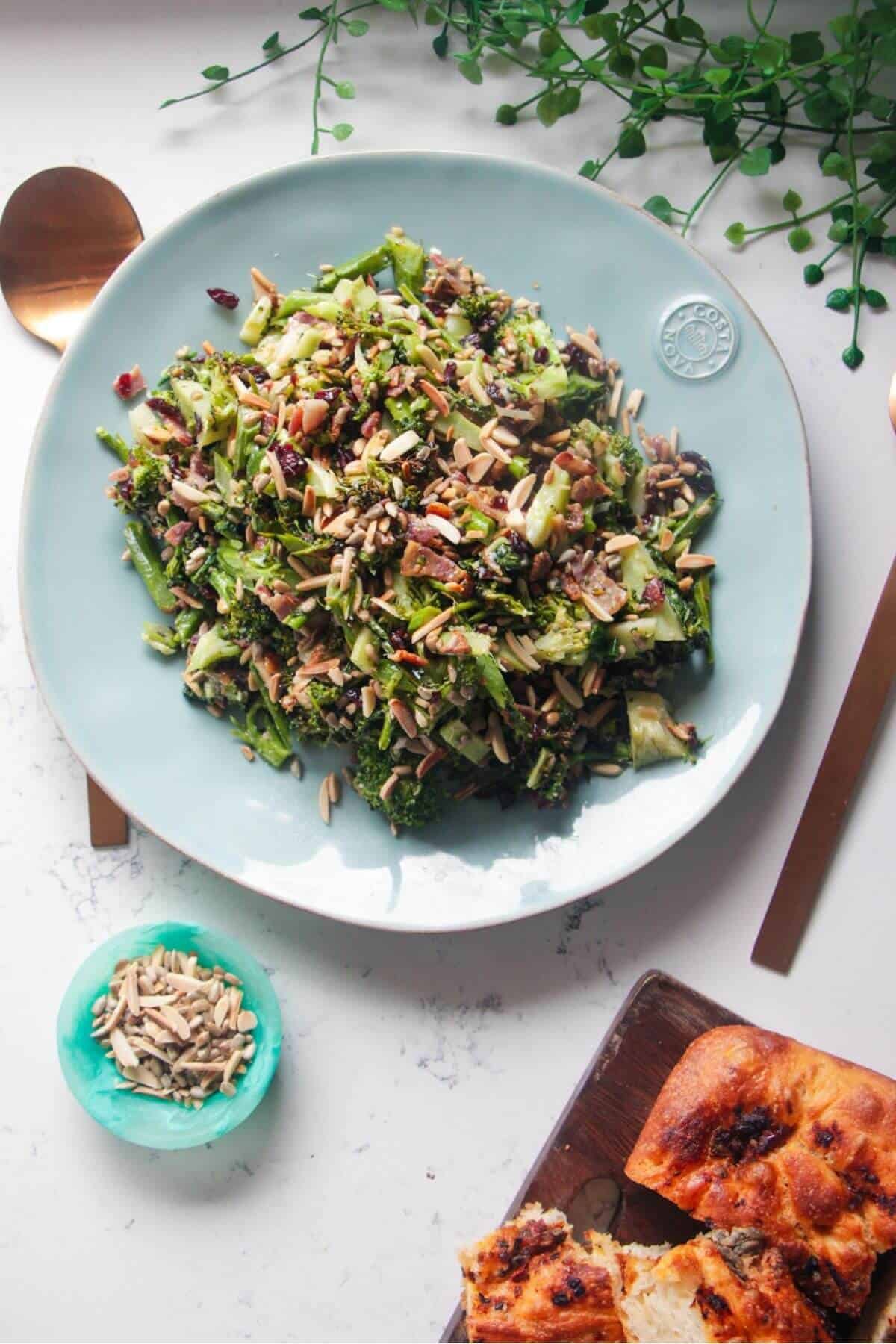 Grilled broccoli crunch salad in a large blue serving plate with focaccia and sunflower seeds on the side.