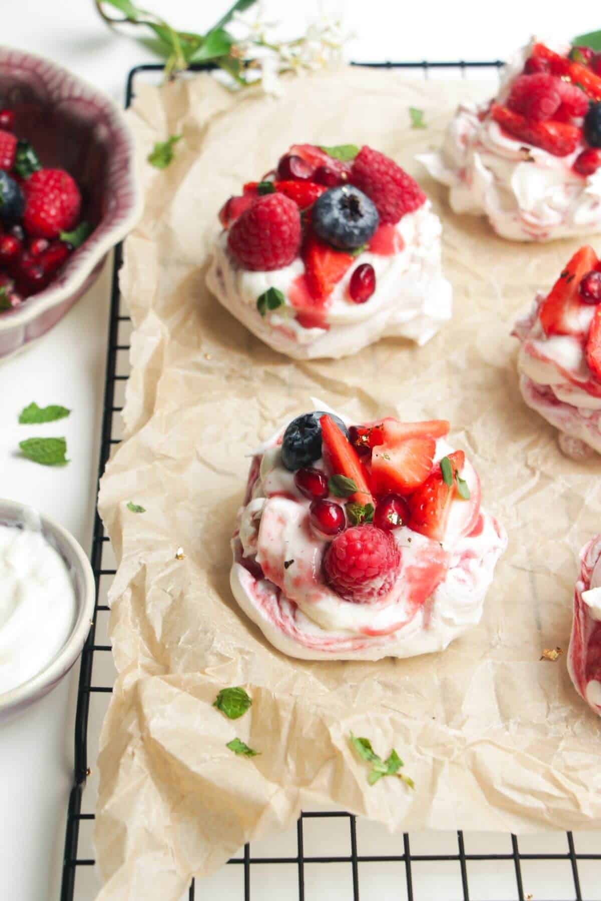 Two mini, berry topped pavlovas on a baking paper lined wire rack with more pavlovas behind them.