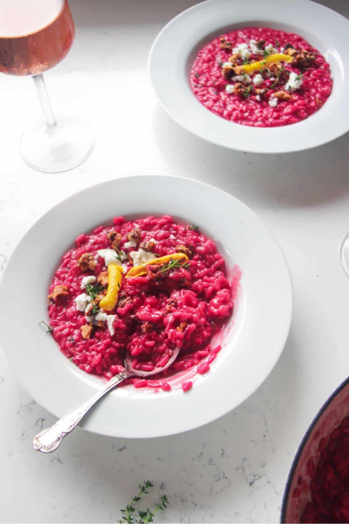 Bright pink beetroot risotto in a white bowl, with feta, lemon peel and walnuts on top with a silver spoon.