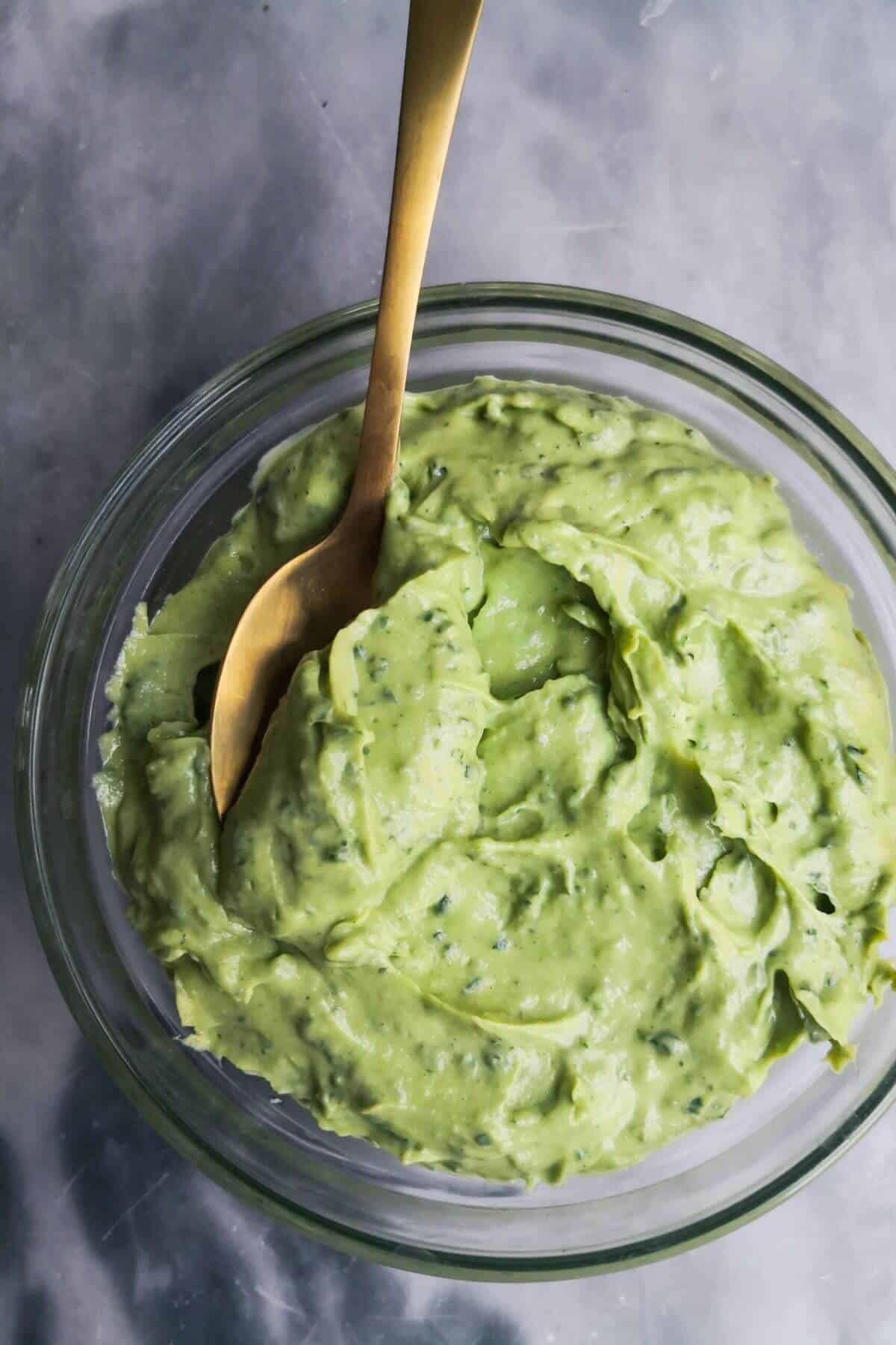 Green tahini in a small glass bowl with a gold spoon in it, on a grey marble background.