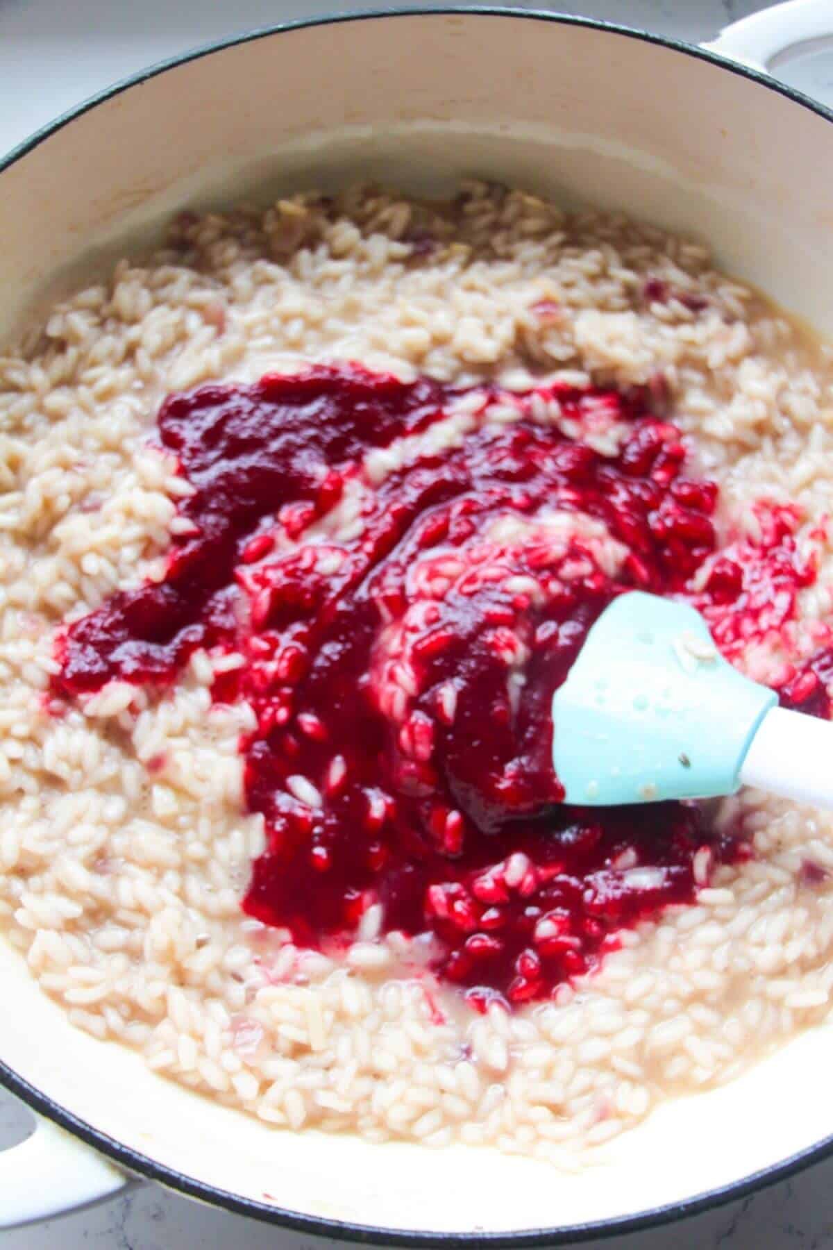 Pink beetroot puree being added to al dente risotto in a large white skillet.