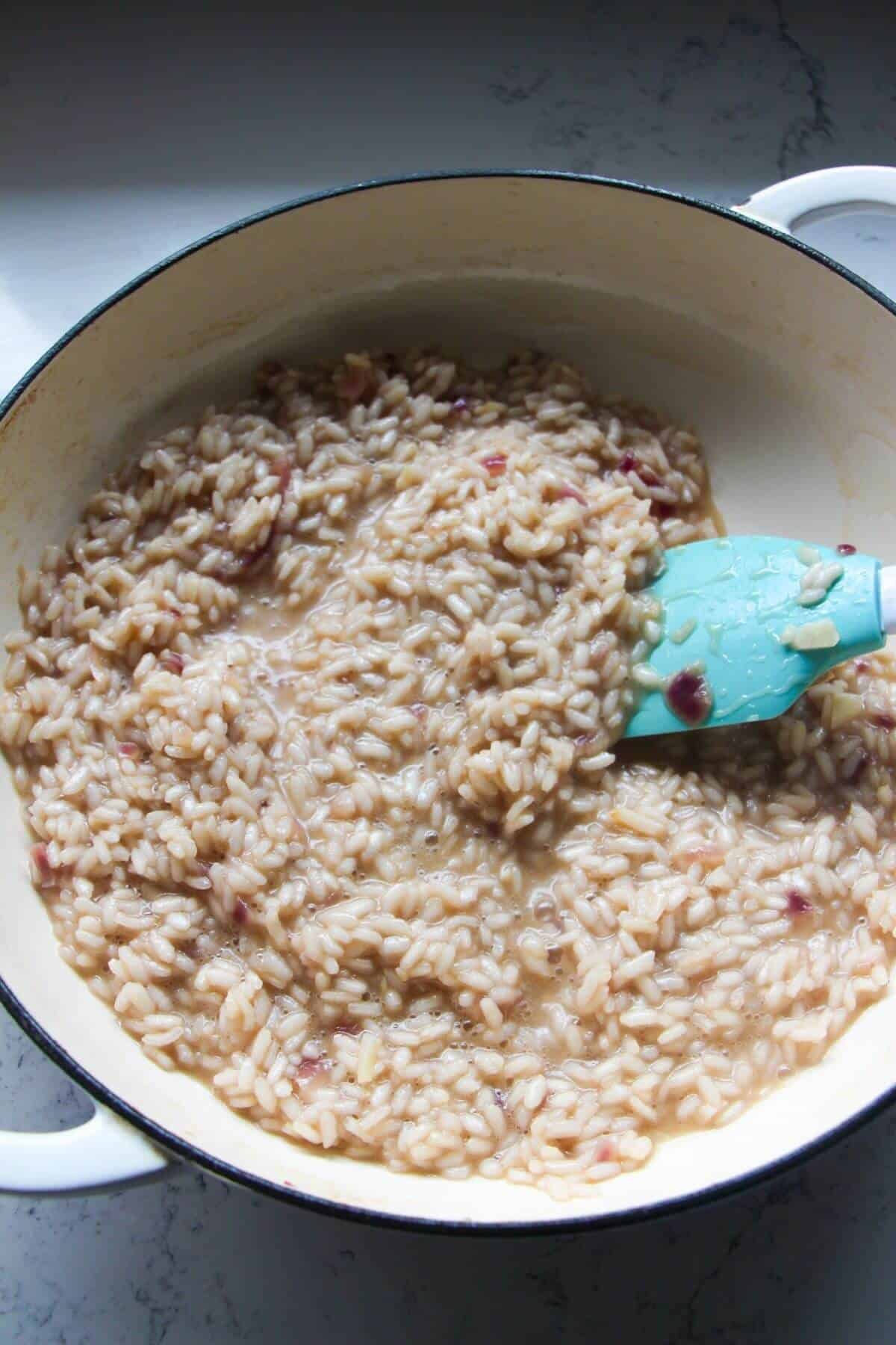 Blue and white spatula stirring al dente risotto in a large white skillet.