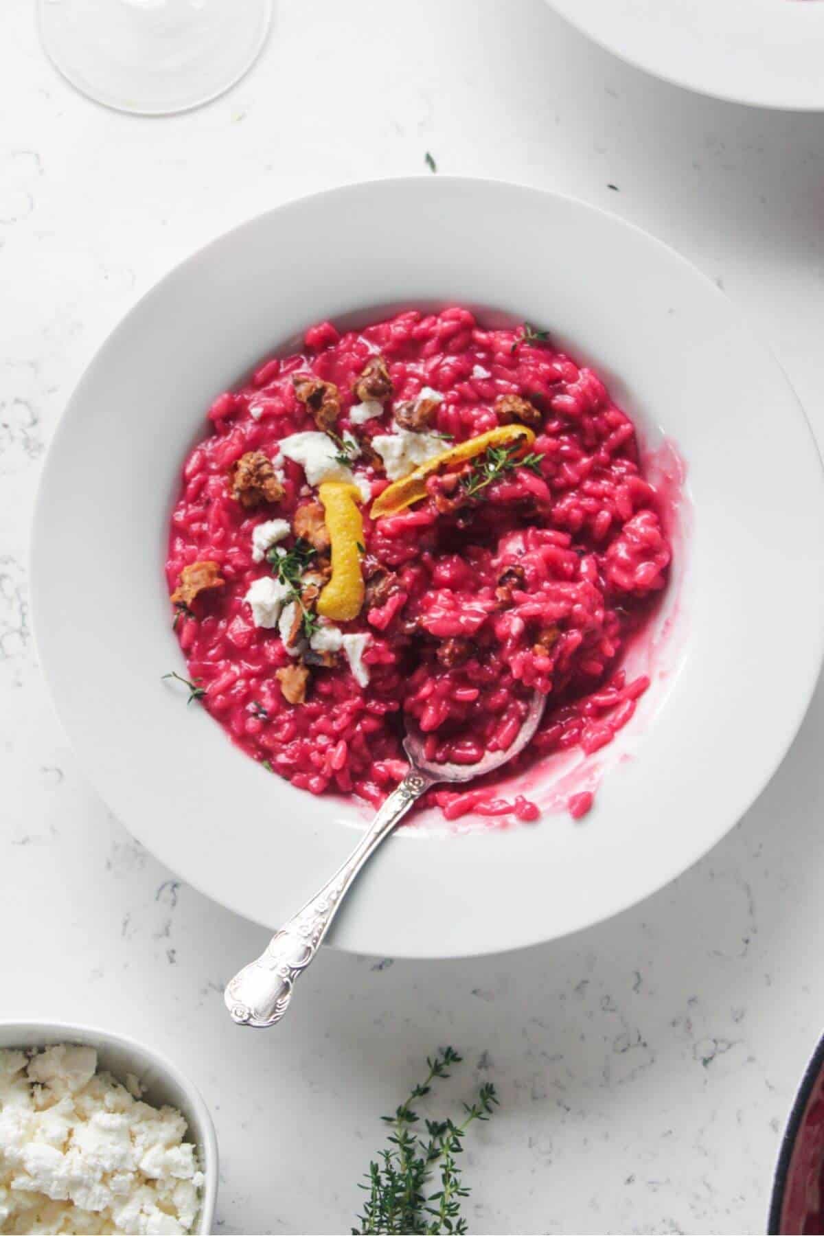 Bright pink beetroot risotto in a white bowl, with feta, lemon peel and walnuts on top with a silver spoon.