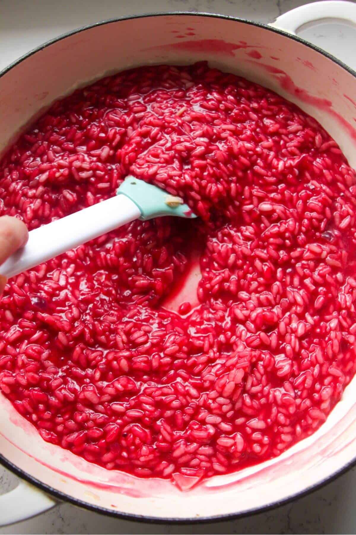 Blue and white spatula stirring bright pink beetroot risotto in a large white skillet.