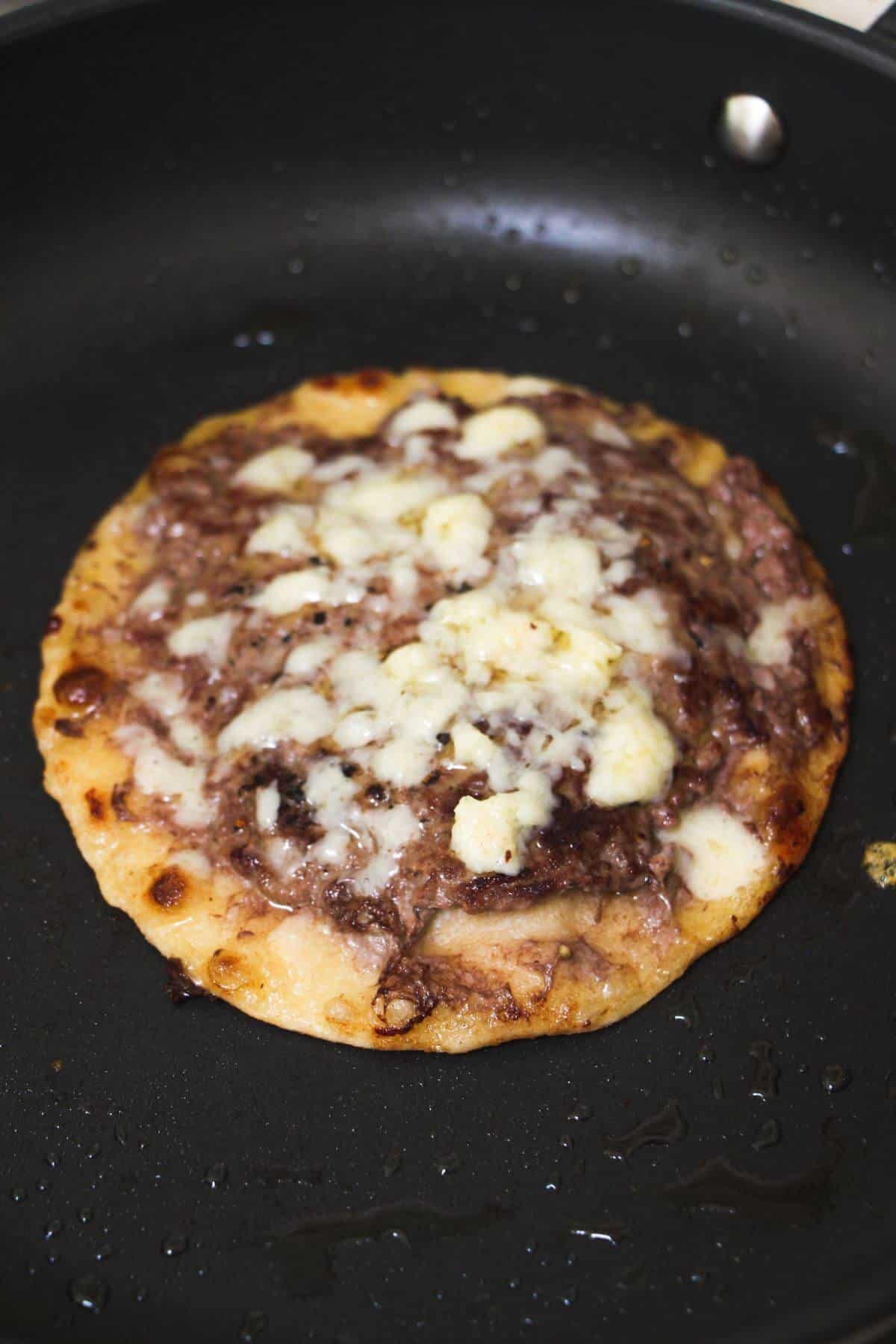 Melted cheese on top of ground beef on a small flour tortilla in a pan.