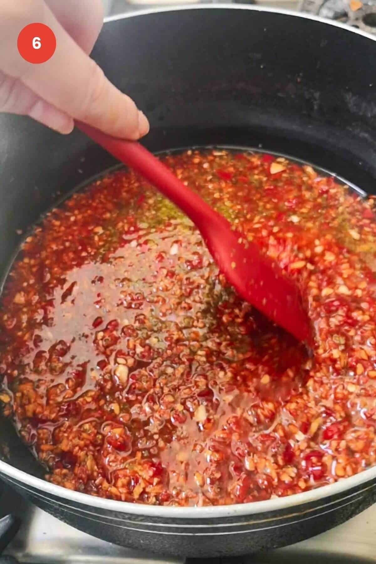 Red spatula stirring chili jam in a large pot.